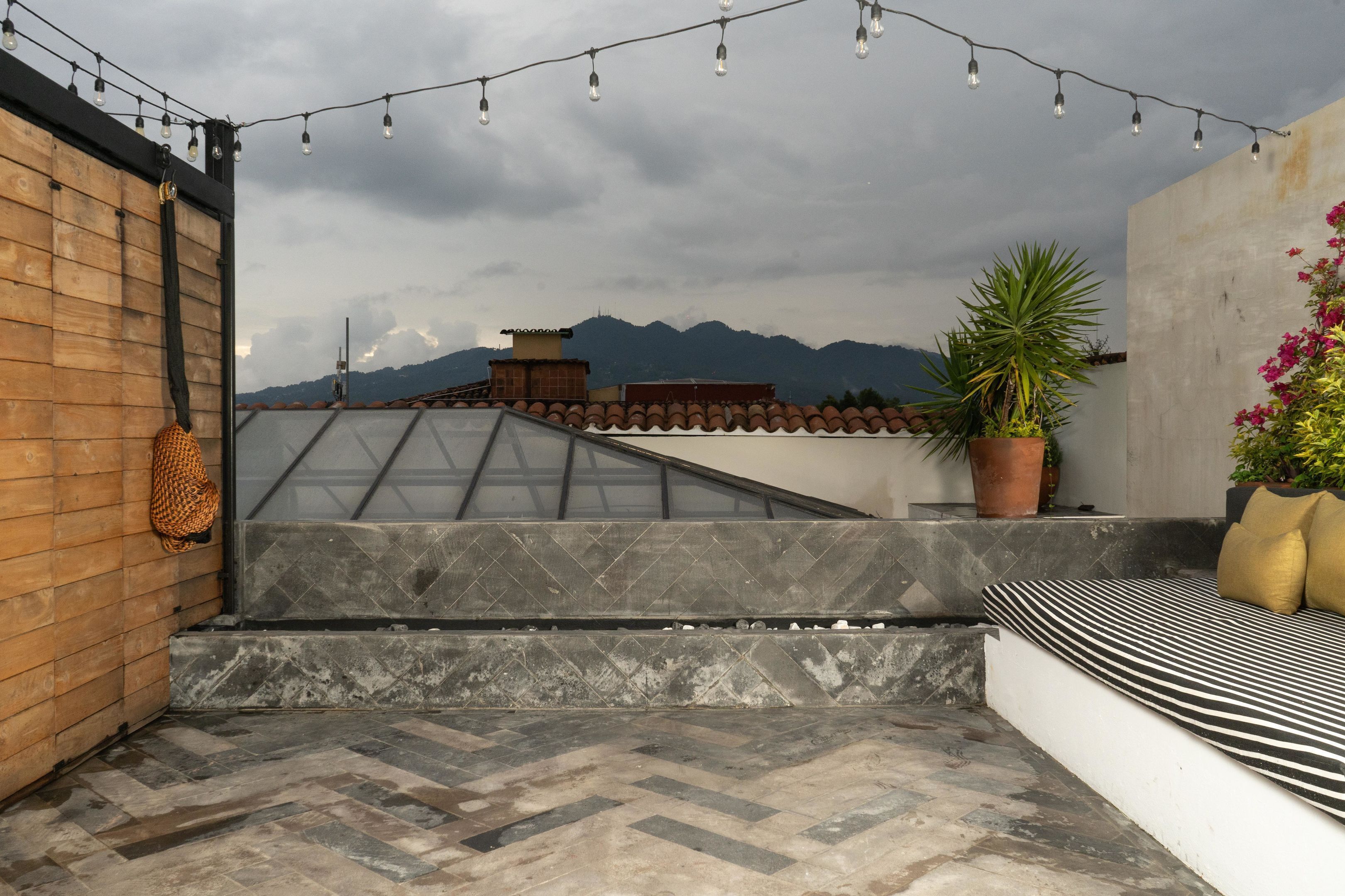 Una terraza en la azotea con suelo de baldosas en espiga, luces colgantes, una planta en maceta y un área de estar con cojines.