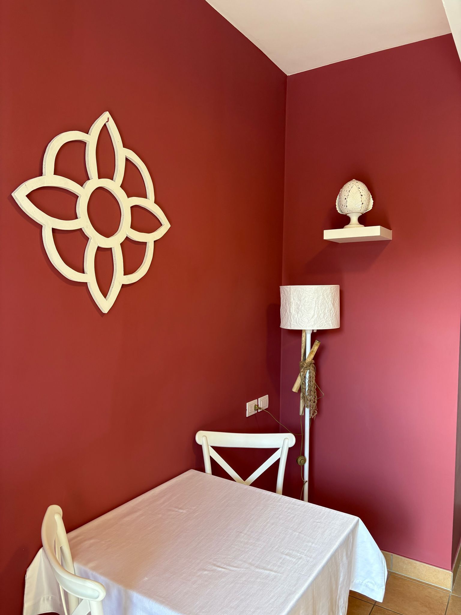 Una habitación con una pared roja, una mesa blanca, una silla blanca, un adorno en la pared, una lámpara y un pequeño estante con un objeto.