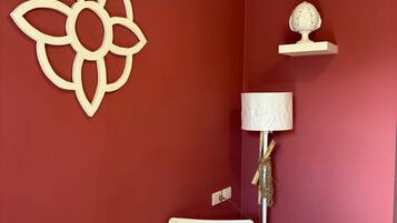 Una habitación con una pared roja, una mesa blanca, una silla blanca, un adorno en la pared, una lámpara y un pequeño estante con un objeto.