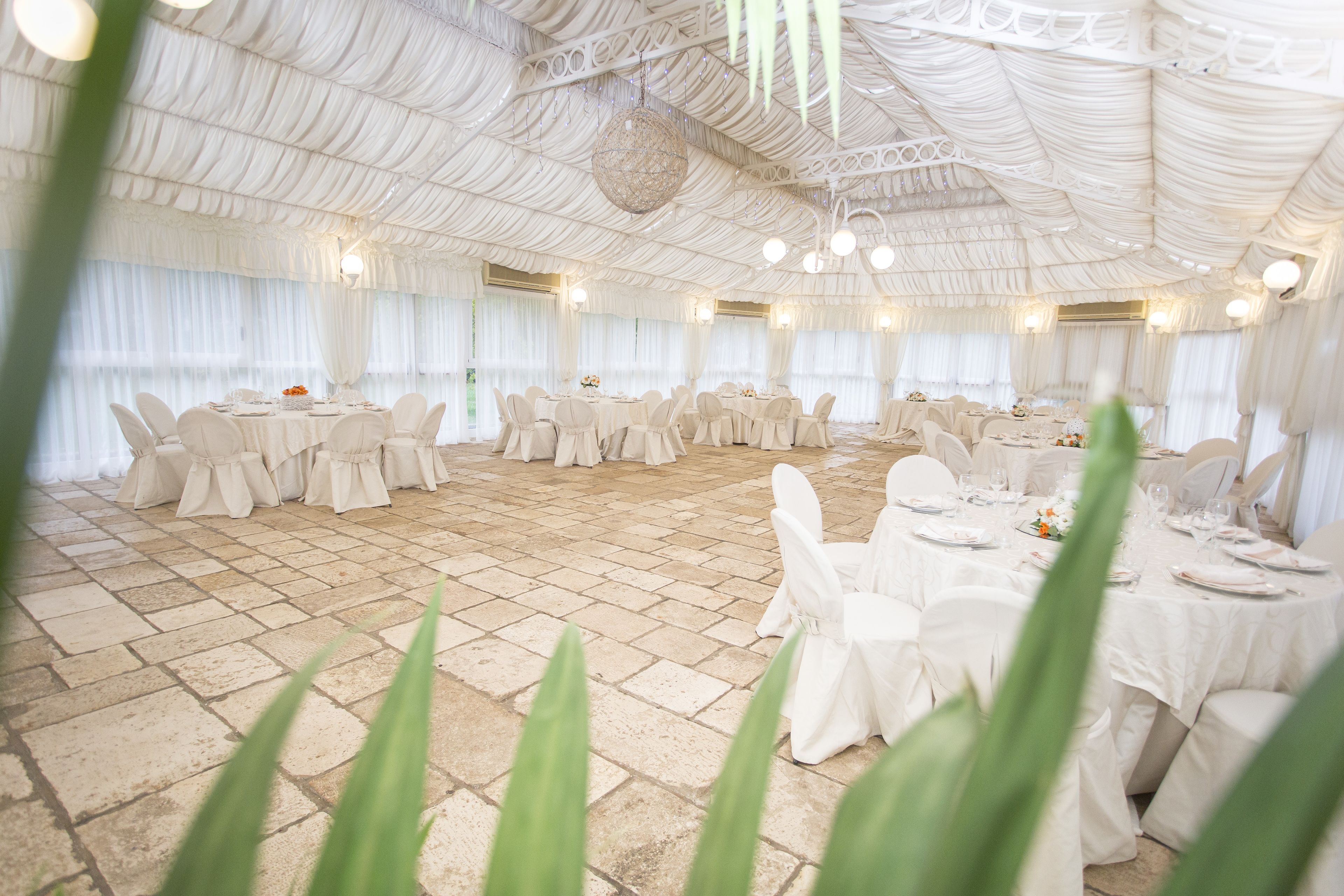 Une salle de banquet avec des tables rondes et des chaises blanches, un sol en pierre et un plafond blanc orné de luminaires suspendus.
