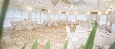 Un salón de banquetes con mesas redondas y sillas blancas, suelo de piedra y techo blanco con luces colgantes.