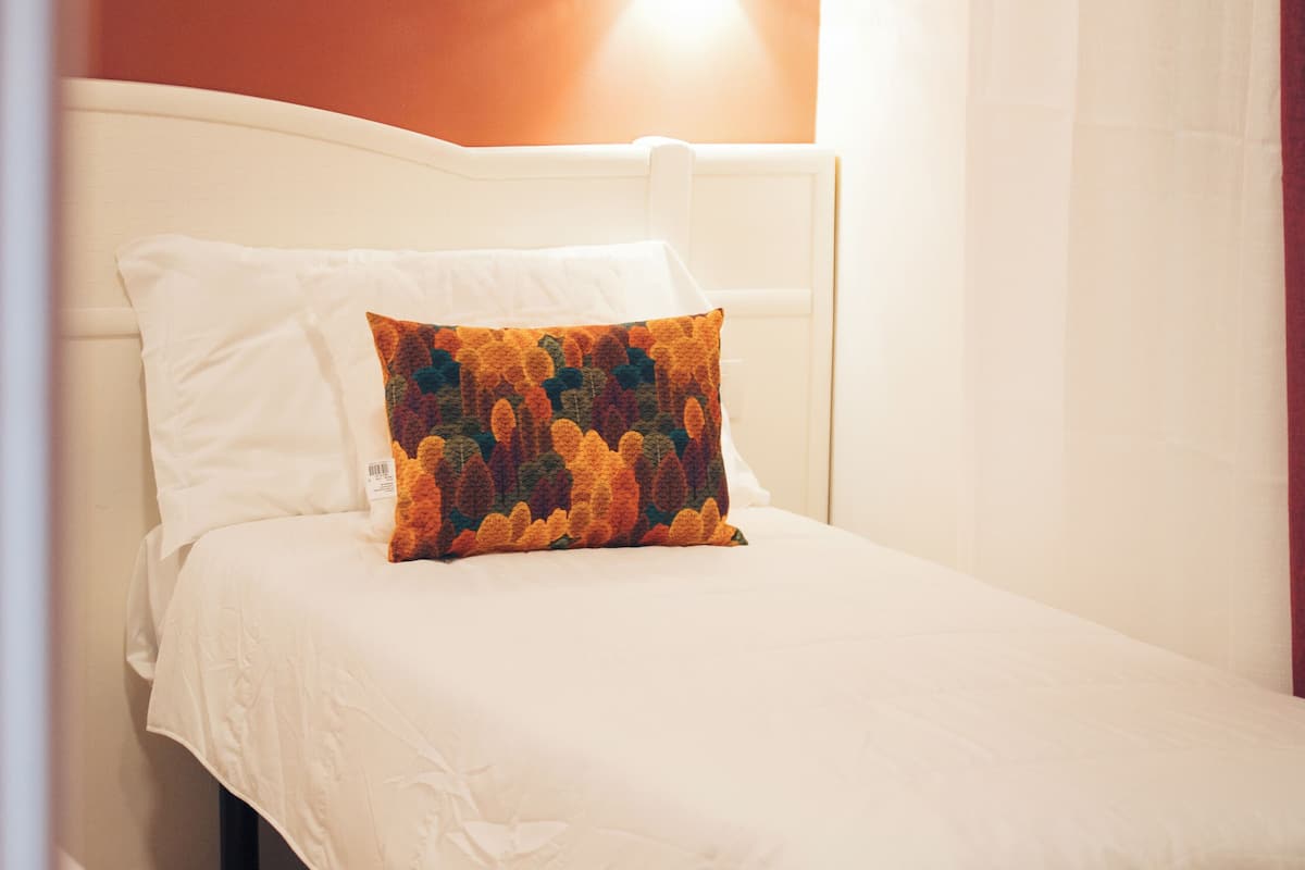 A neatly made bed with white bedding and a colorful pillow, a wall-mounted light fixture, and a curtain partially drawn to the side.