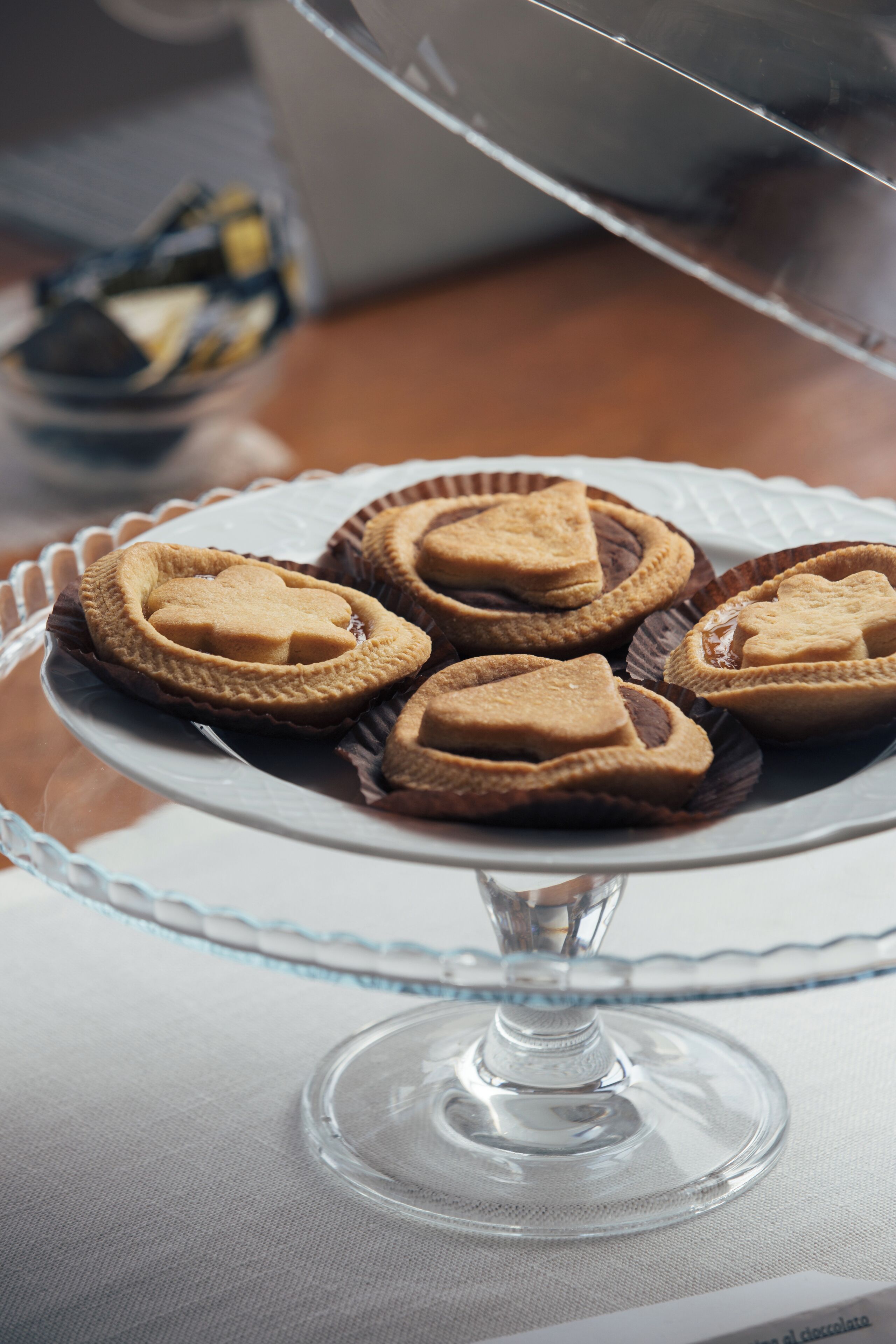 A clear glass cake stand with a white base and a scalloped edge, holding several heart-shaped cookies in brown paper cups.