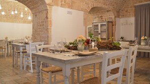 Restaurante com paredes de pedra e tetos abobadados, mesas e cadeiras de madeira branca, e decoração rústica com flores e uma gaiola de pássaros.