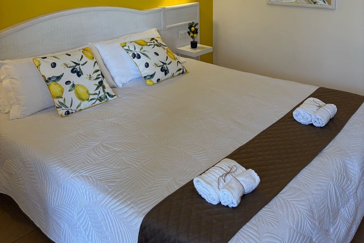 A neatly made bed with white linens and decorative pillows, a bedside table with a lamp, and a large flower wall art piece.