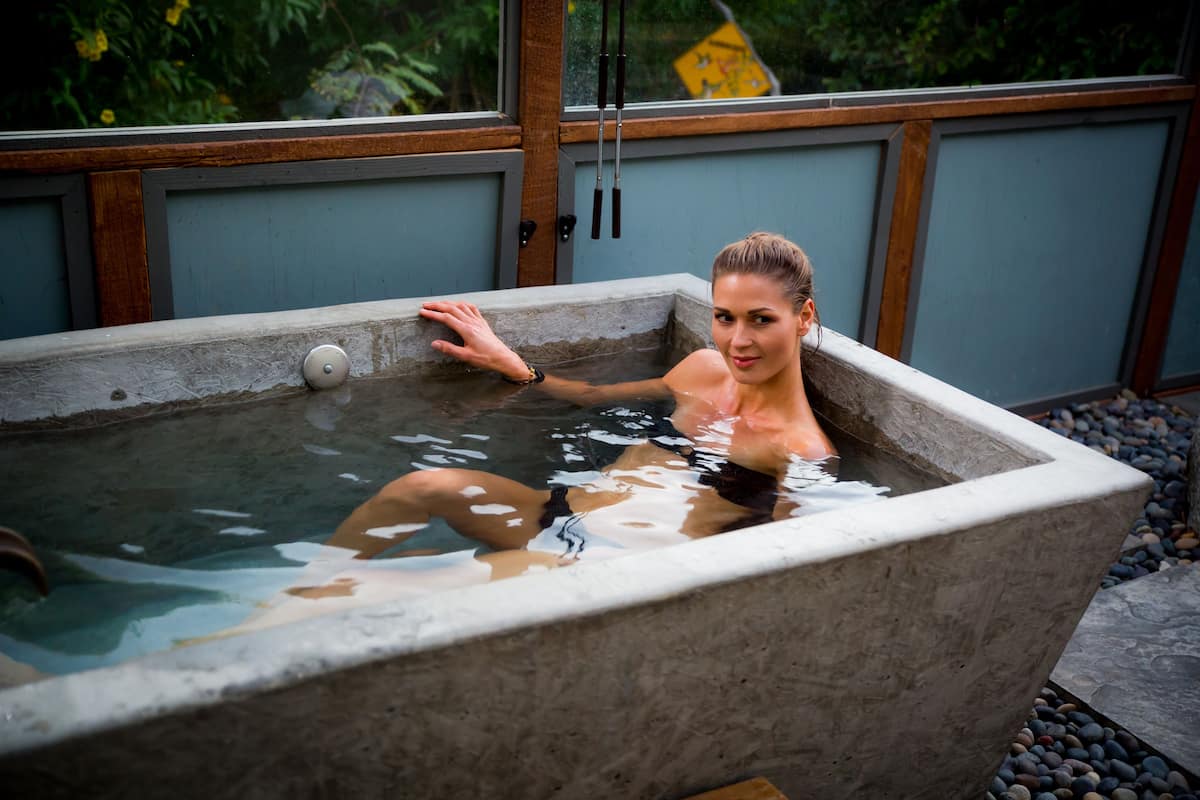 A person in a stone hot tub with a wooden frame and a window in the background.