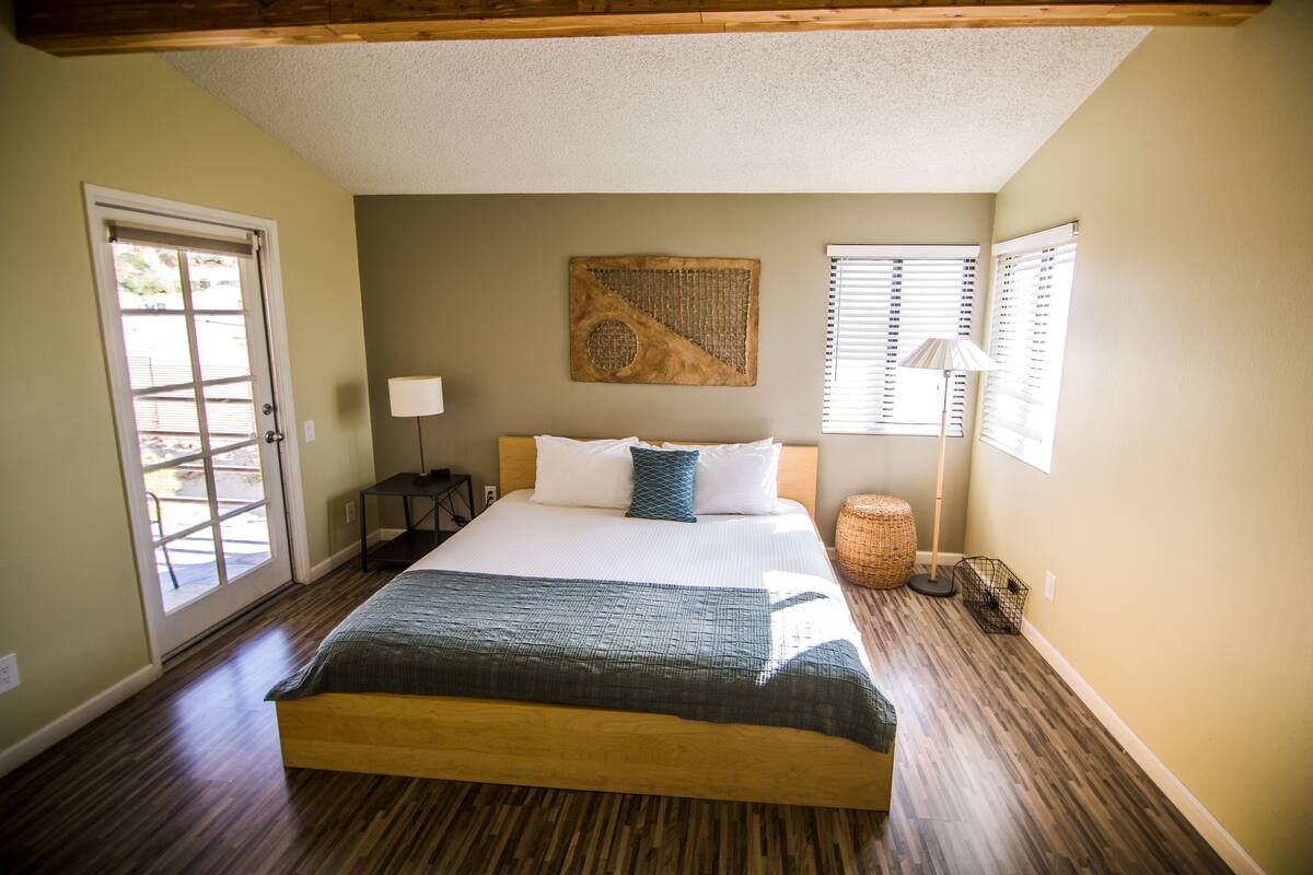 A bedroom with a large bed, a door to a patio, a wall-mounted artwork, and a window with blinds.