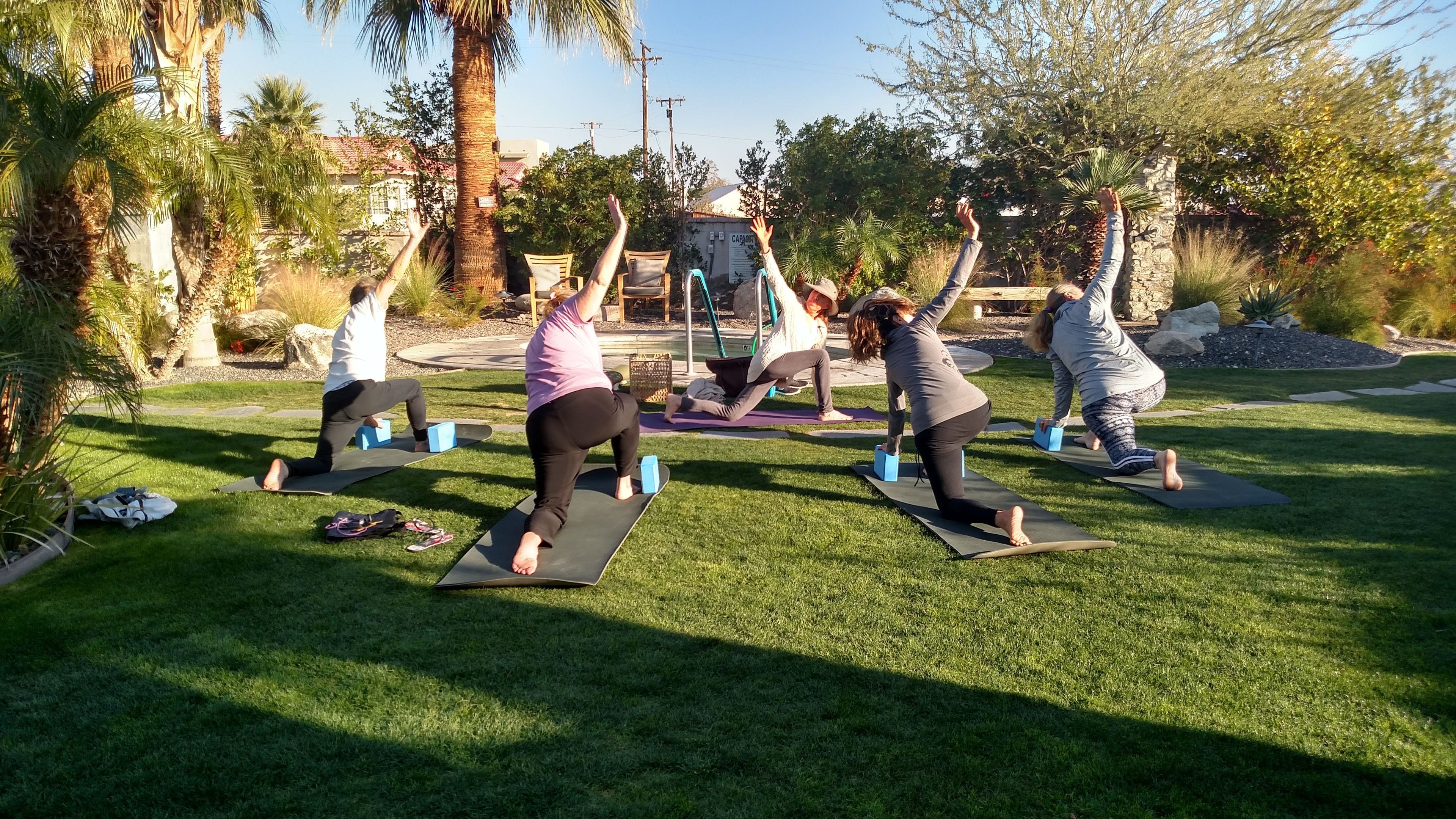 A group of people performing yoga outdoors on mats with trees and a building in the background.