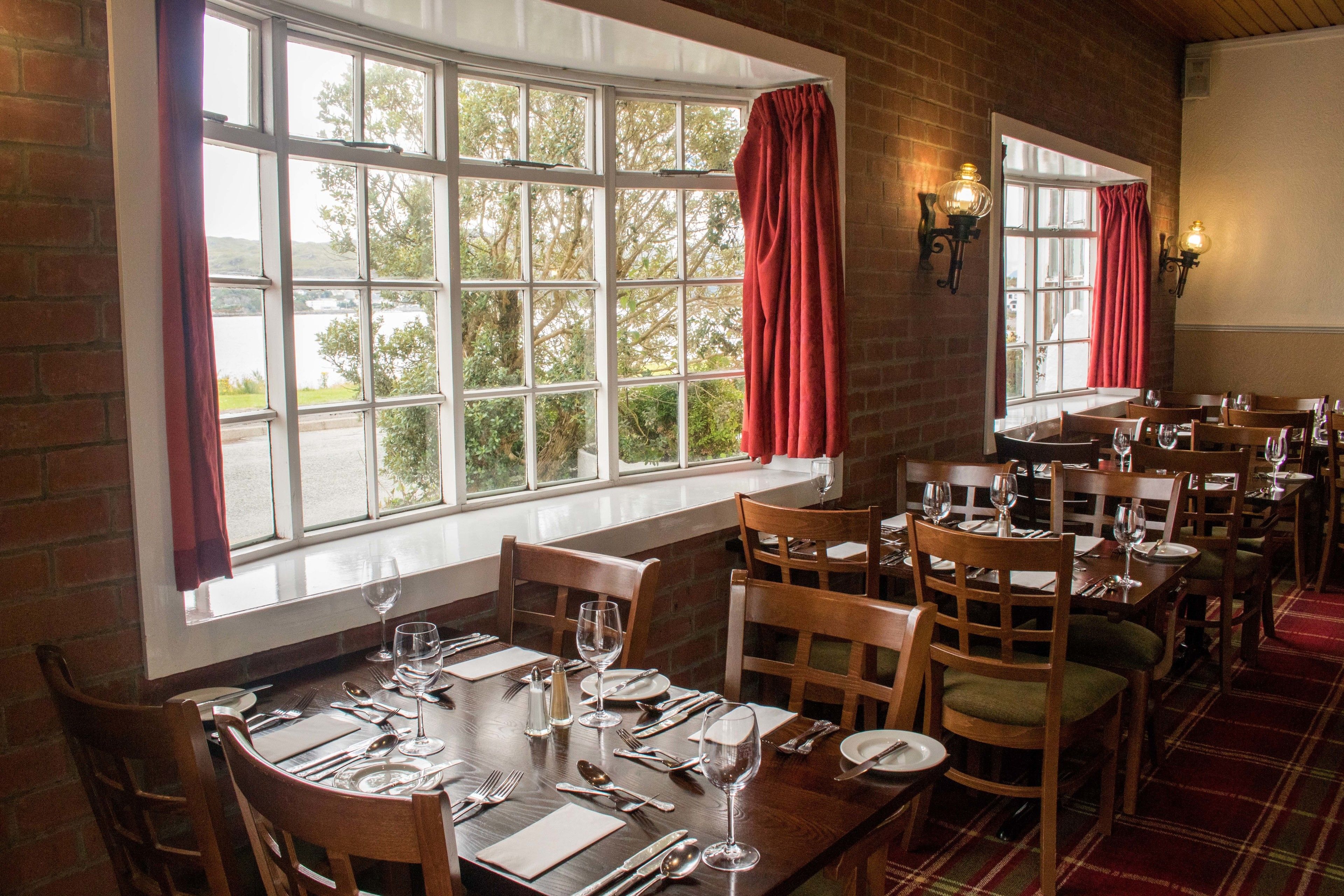A restaurant with multiple tables set for dining, large windows with curtains, and brick walls.