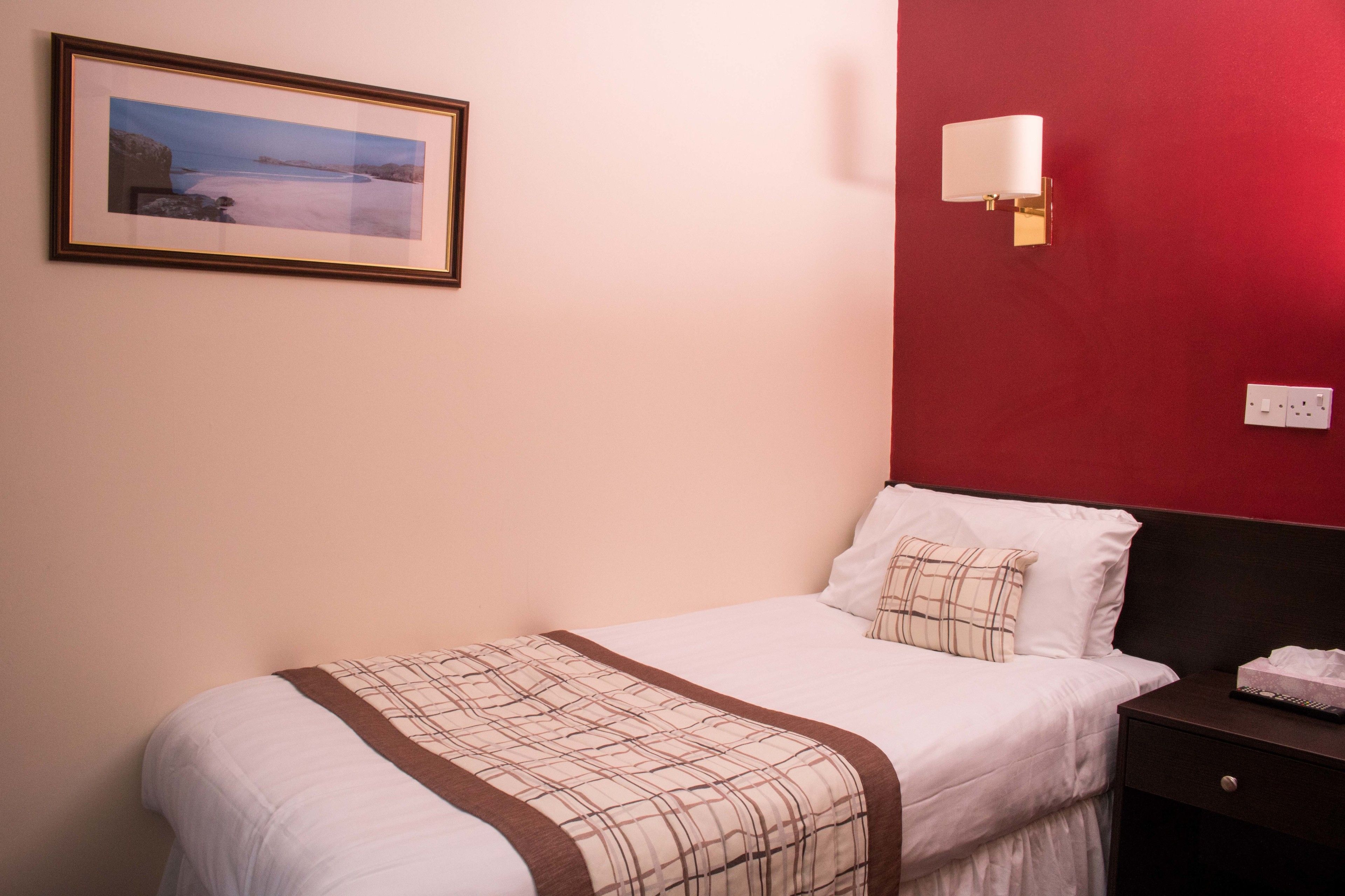 Una cama bien hecha con una manta escocesa, una mesita de noche con un control remoto y una lámpara de pared.