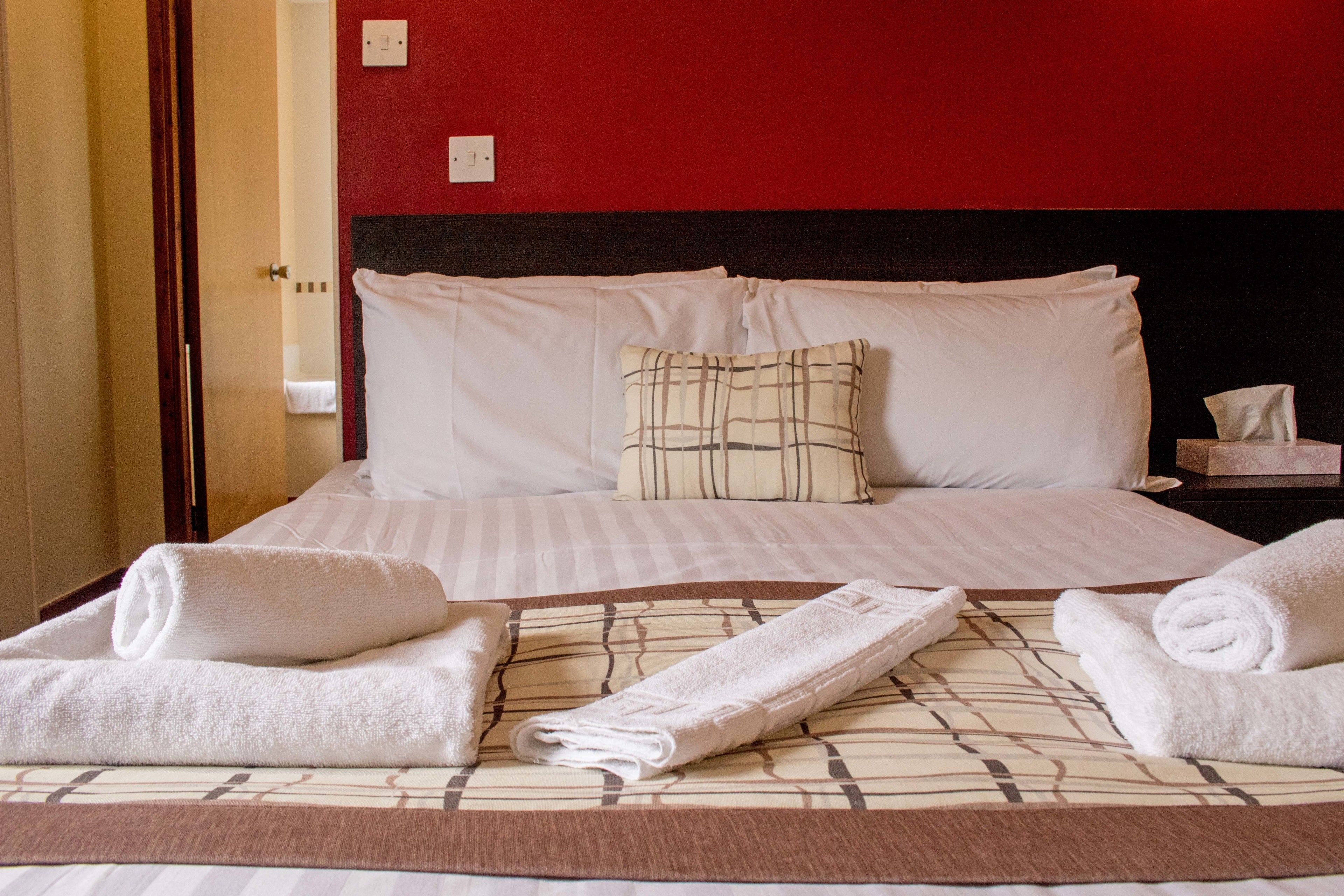 Un lit bien fait, avec des draps blancs et un coussin à carreaux, une table de chevet avec une boîte de mouchoirs et une serviette enroulée posée sur le lit.