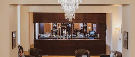 A bar area with a chandelier, wooden cabinets, and a bar counter with various bottles. There are seating areas with chairs and a table.