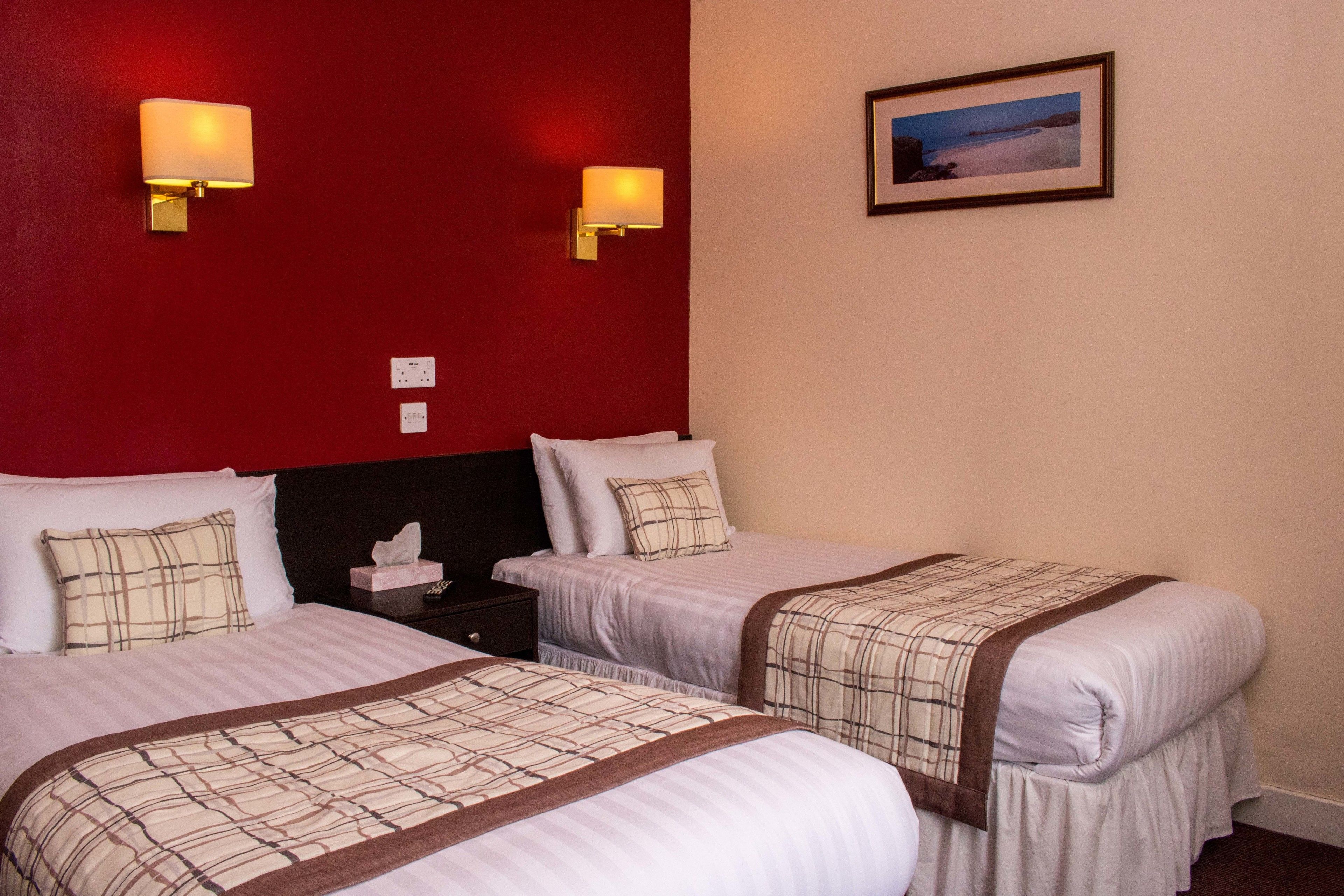 A hotel room with two beds, each with a white bedspread and brown trim, a nightstand with a lamp, and a framed picture on the wall.