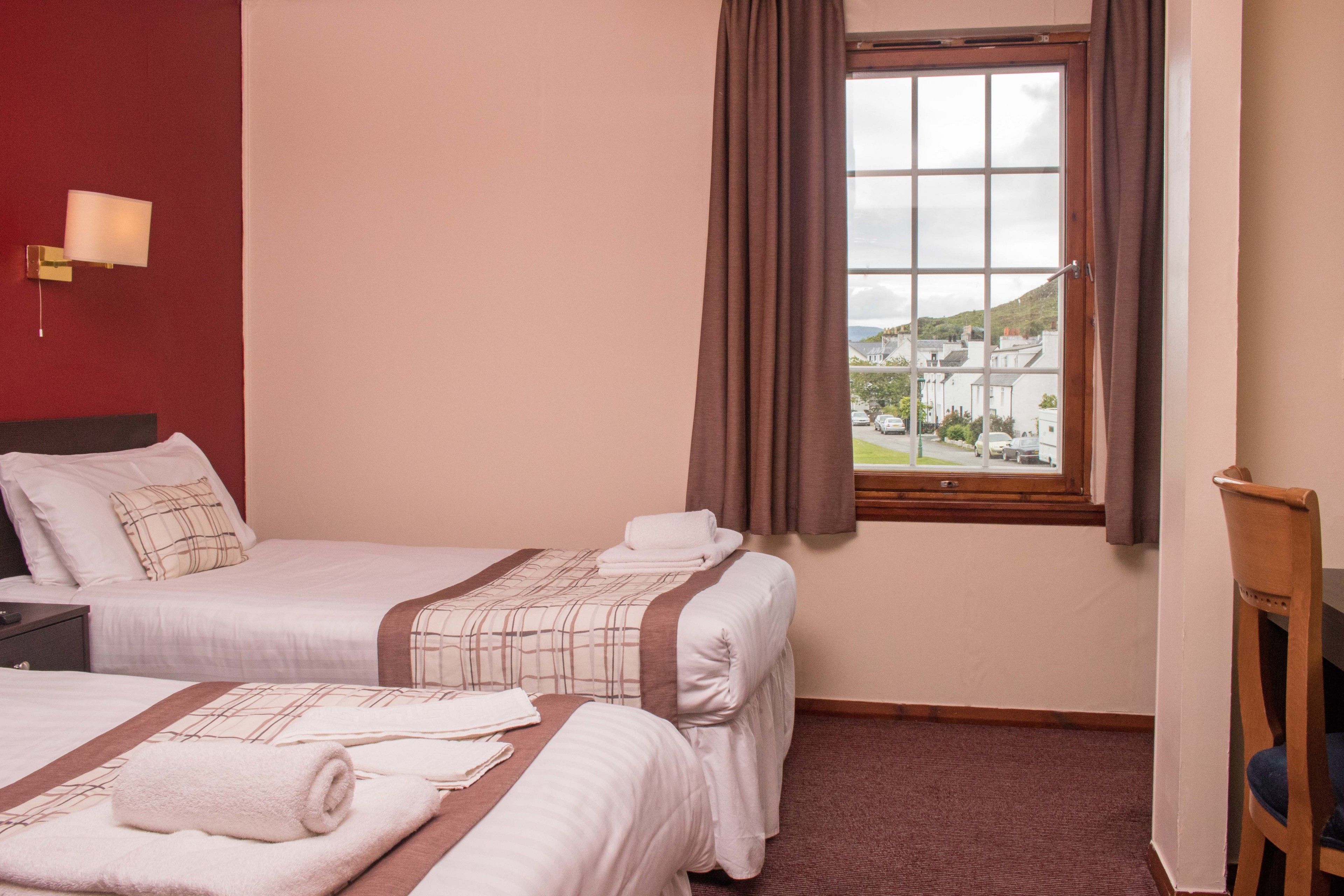 A hotel room with two beds, a window with curtains, a nightstand, and a lamp.