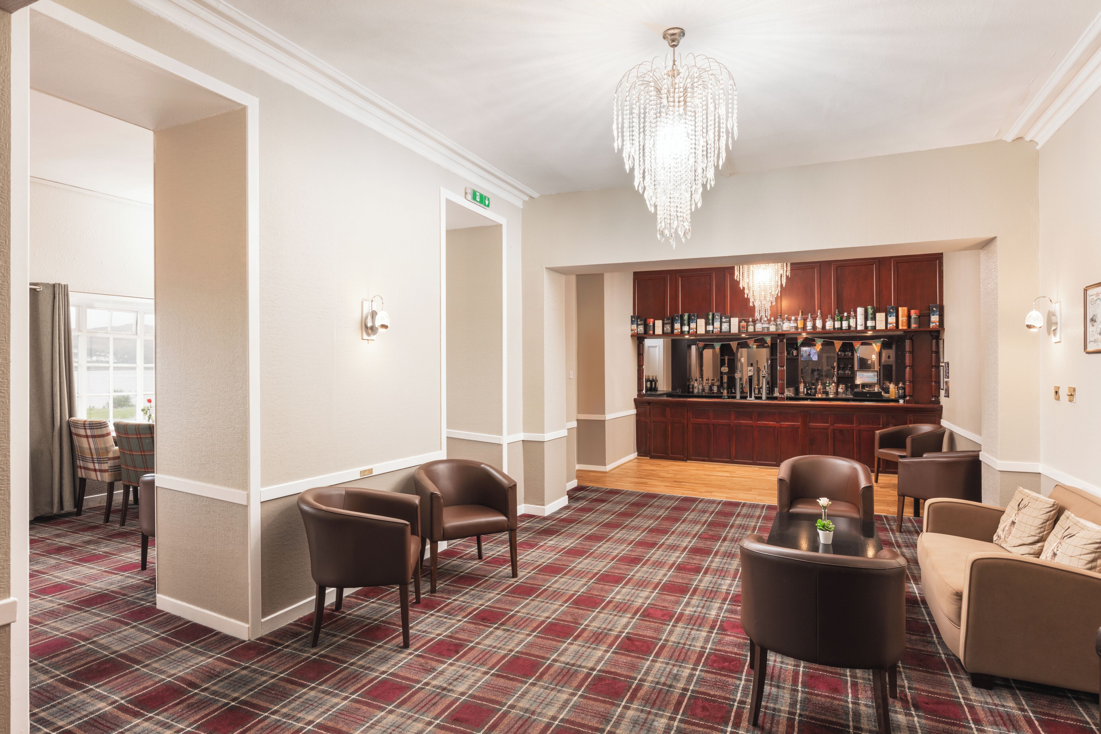 A hotel lobby with a bar area, plaid carpet, leather chairs, and a chandelier.