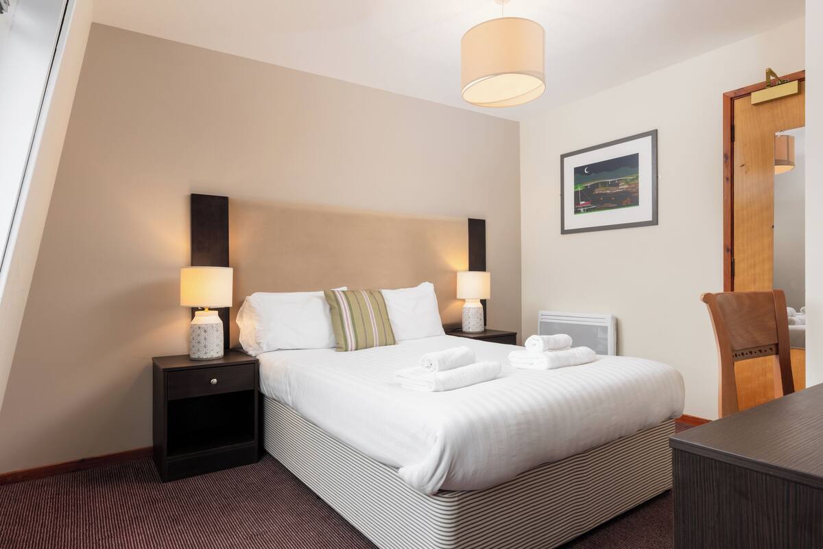 A hotel room with a large bed, two bedside tables with lamps, a wooden chair, a desk, and a framed picture on the wall.