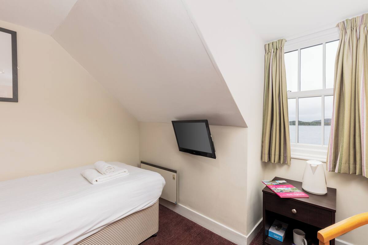 A hotel room with a single bed, a flat-screen TV mounted on the wall, a bedside table with a lamp and a book, and a window with curtains.