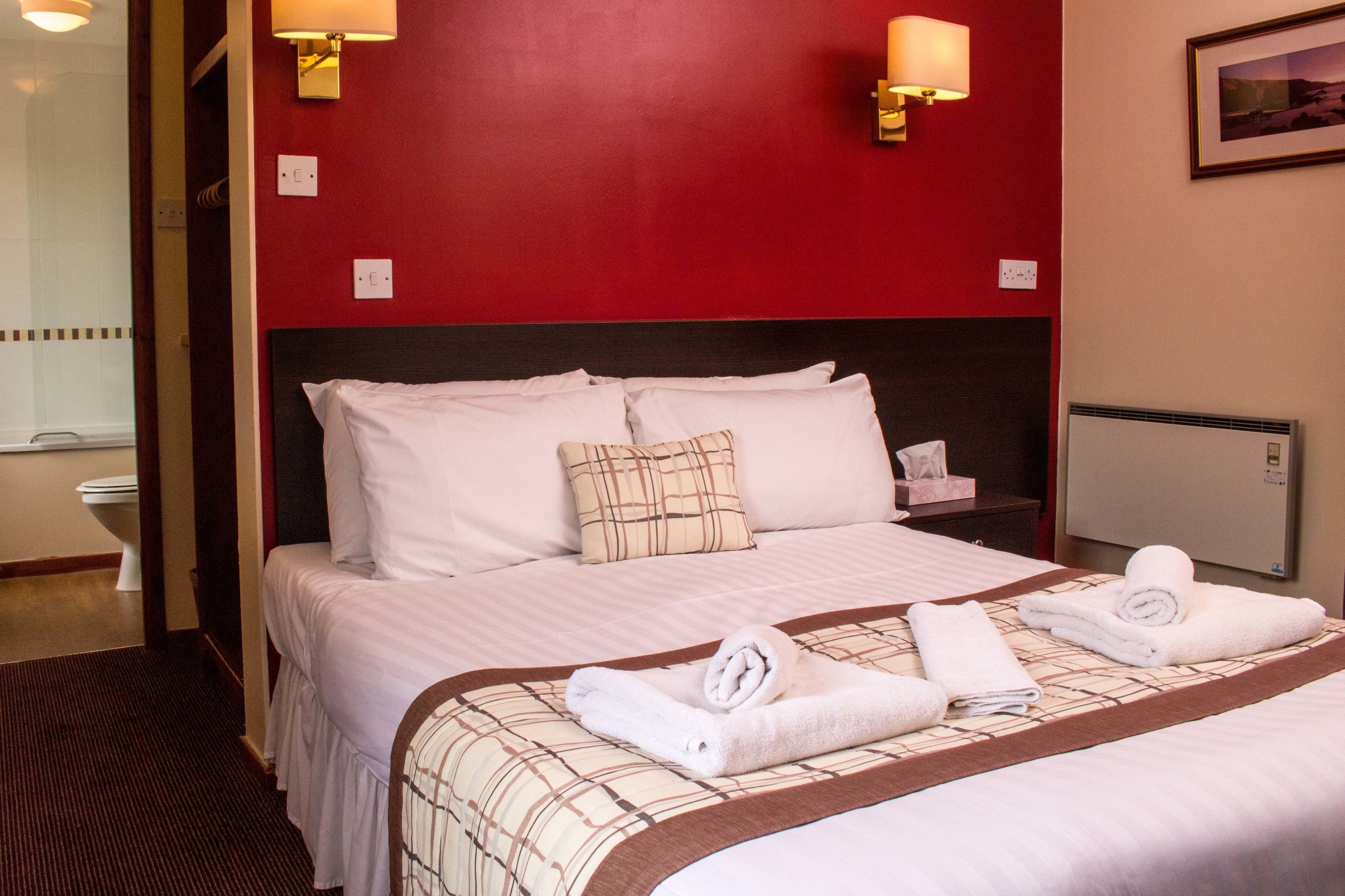 Een hotelkamer met een rode accentmuur, een groot bed met witte beddengoed, twee wandlampen en een ingelijst schilderij aan de muur.