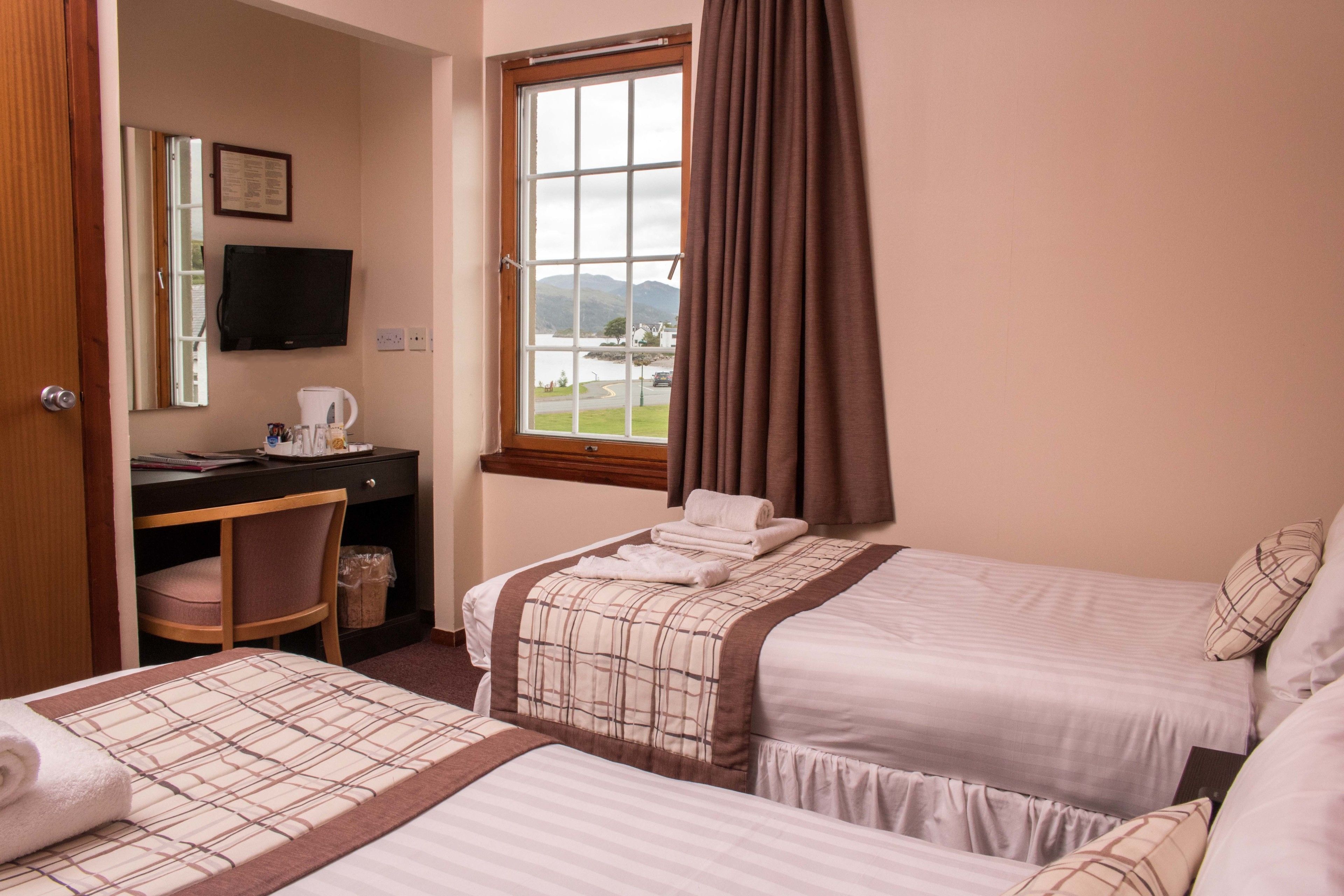 A hotel room with two beds, a desk with a chair, a television, and a window with a view of a lake and mountains.