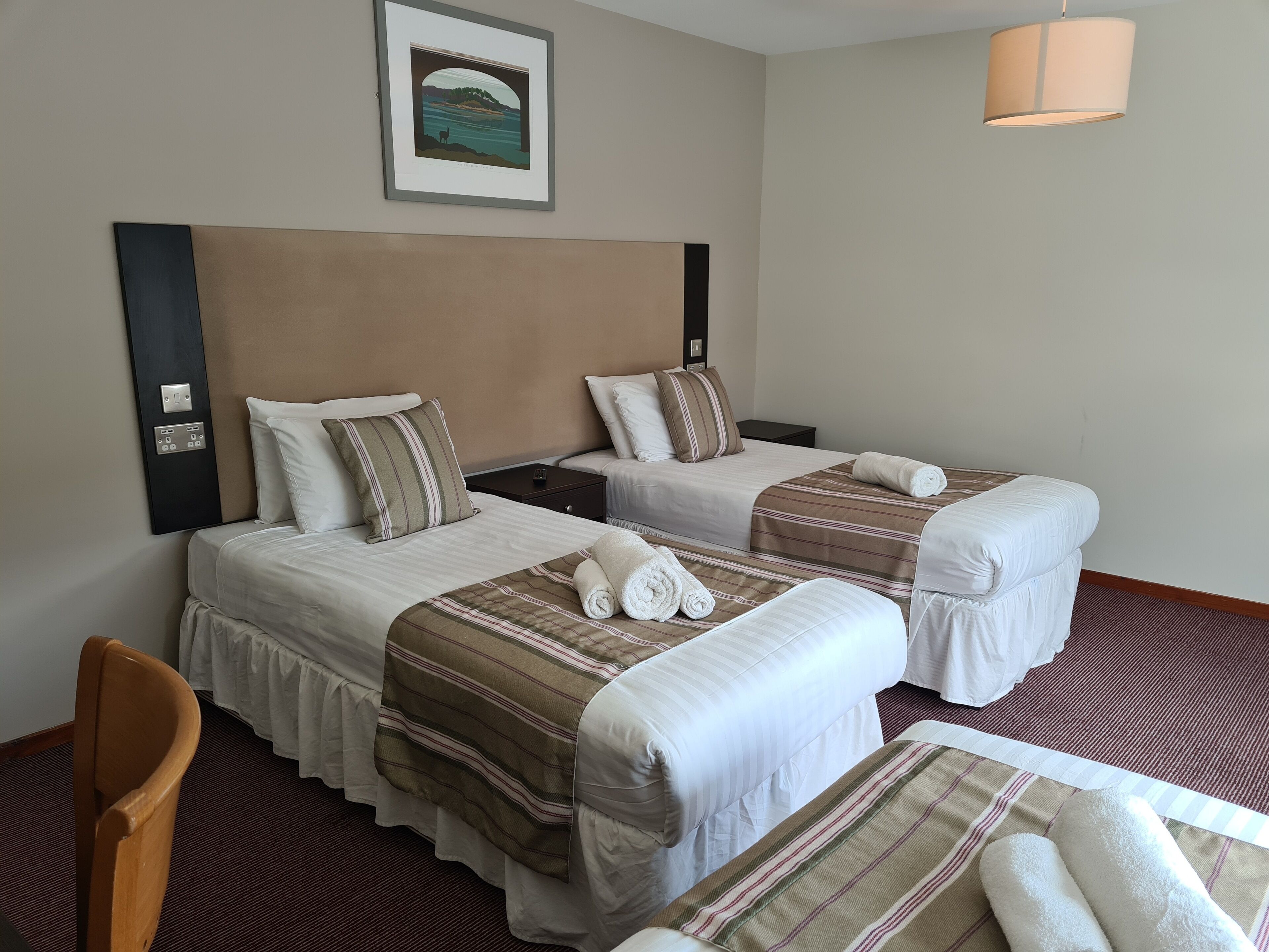 Habitación de hotel con dos camas, cada una con cubrecama a rayas, cabecero y almohada. Se observa un cuadro enmarcado en la pared, una lámpara y una silla.