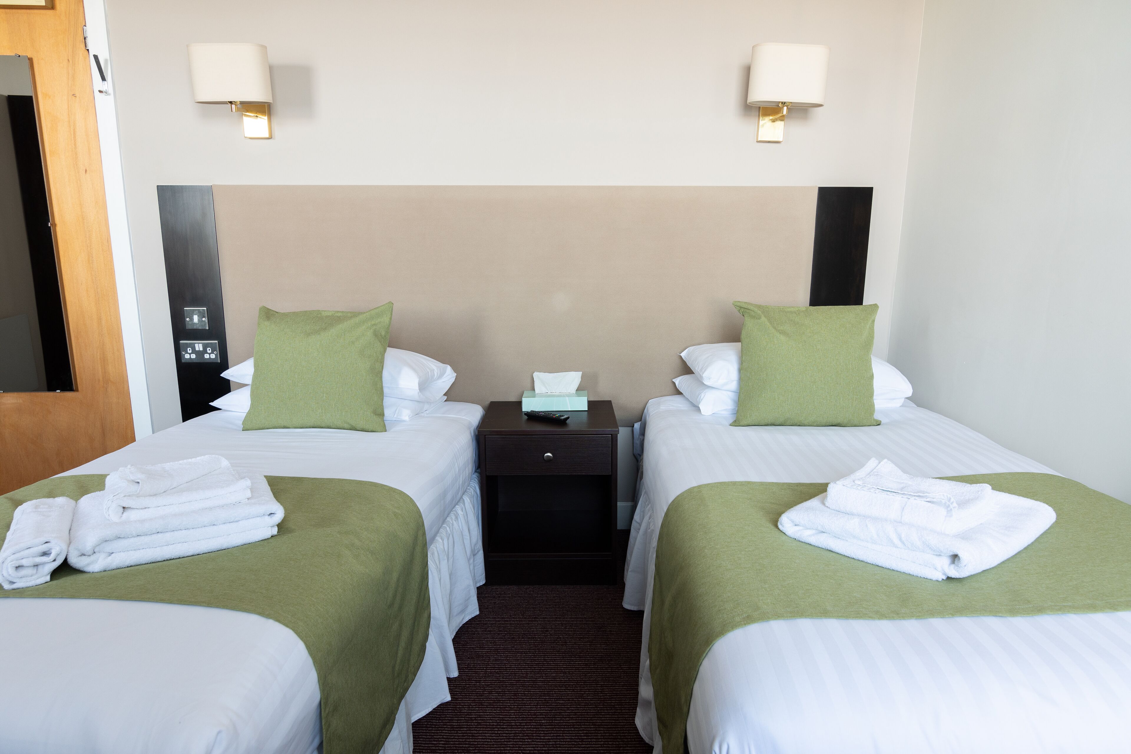 A hotel room with two beds, each with green bedspreads and white linens, a nightstand with a lamp, and a wooden door.