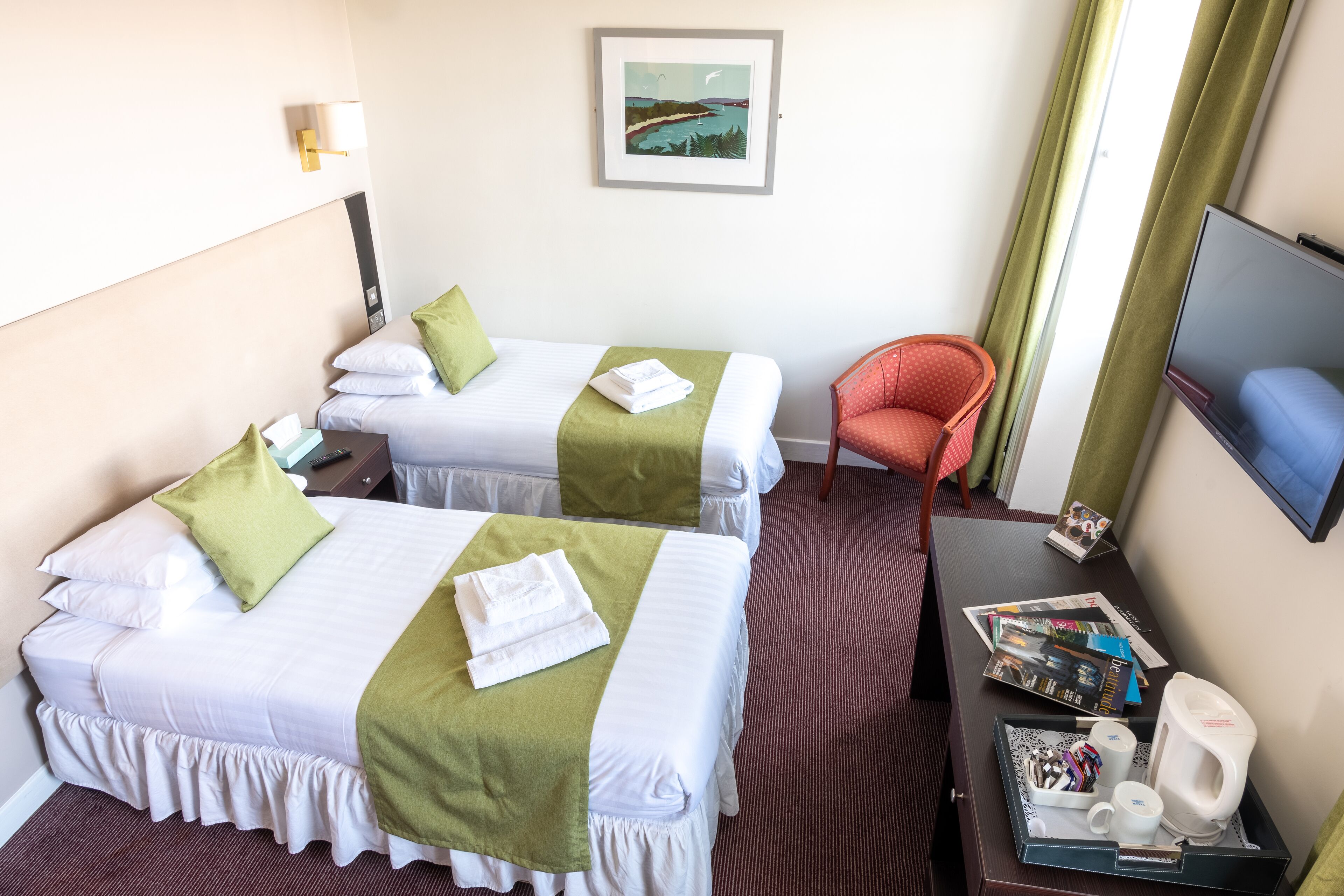 Una habitación de hotel con dos camas, un escritorio con cafetera, revistas y televisión.