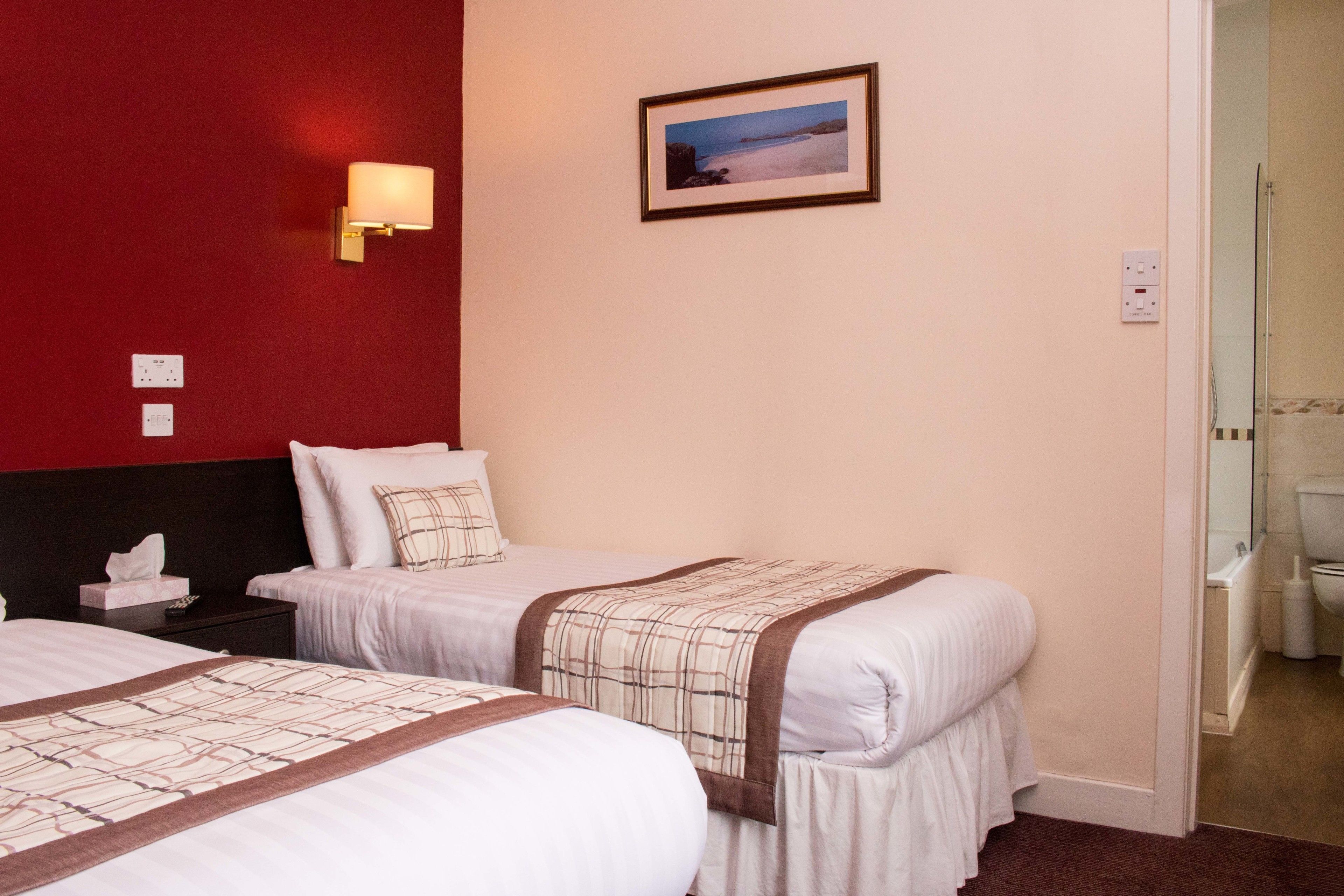 A hotel room with two beds, a red accent wall, a framed picture, and a bathroom visible through an open door.