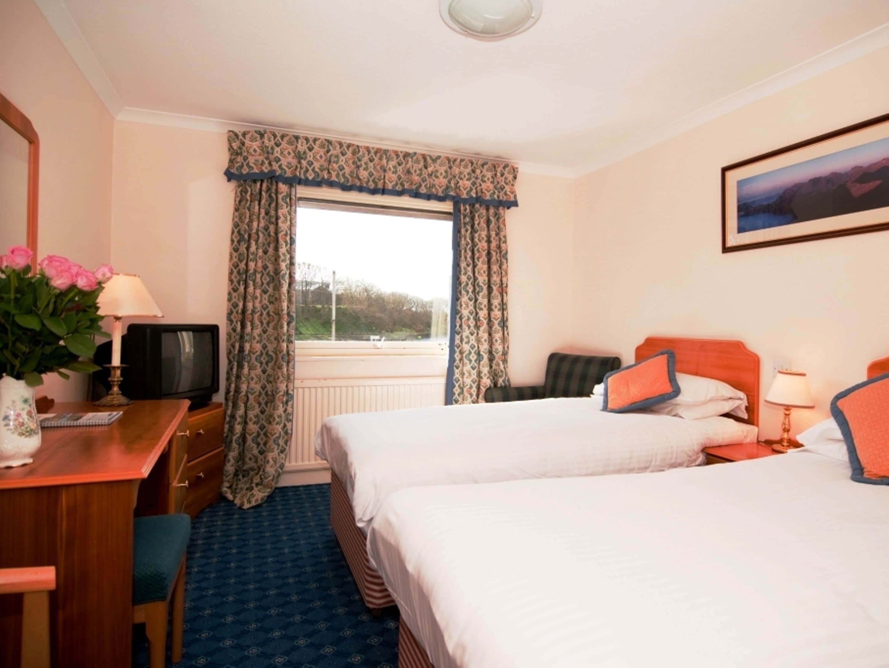 A hotel room with two beds, a desk with a television, a window with curtains, and a painting on the wall.