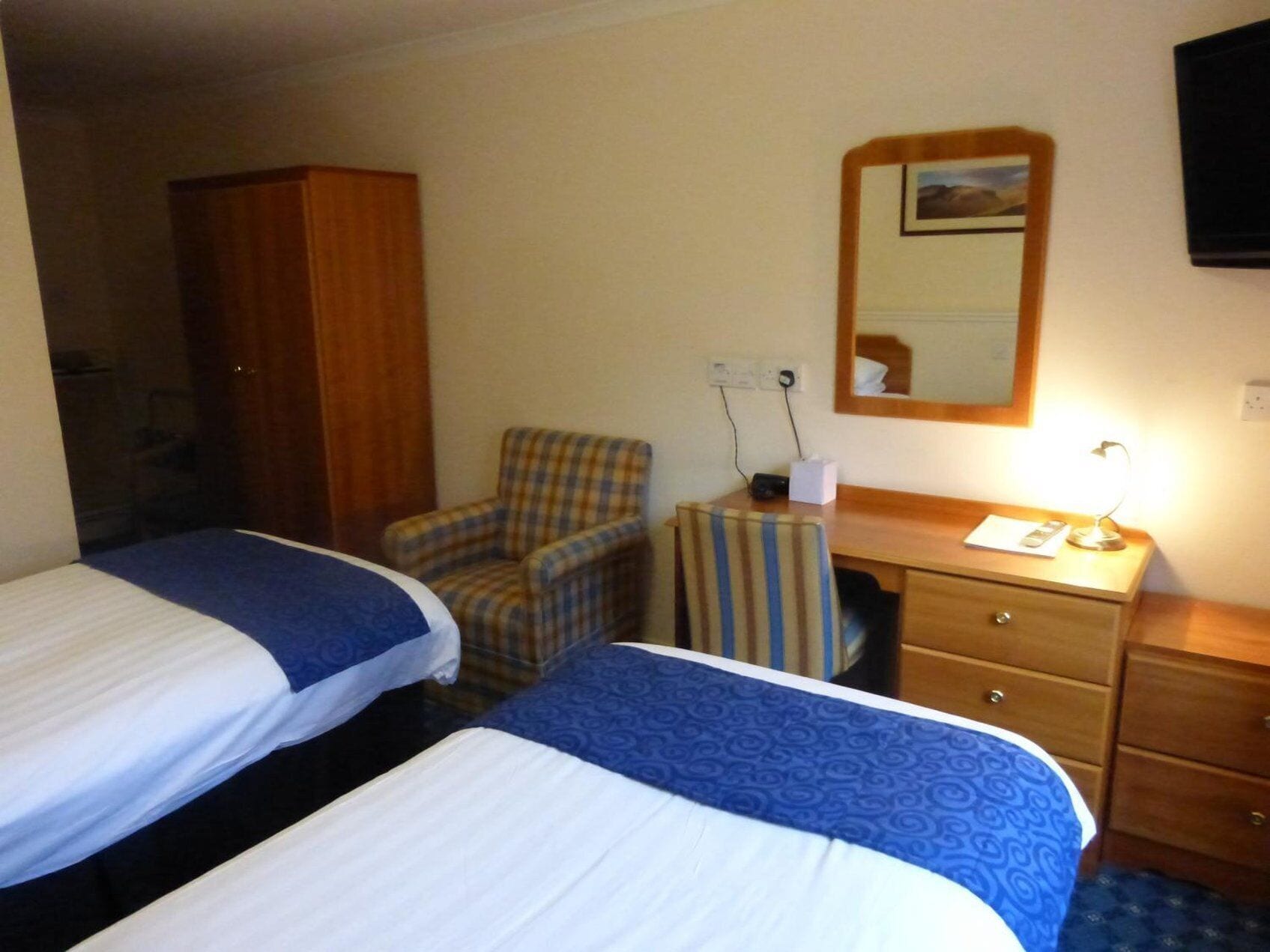 A hotel room with two beds, a wooden wardrobe, a plaid armchair, a small desk with a lamp, and a mirror on the wall.