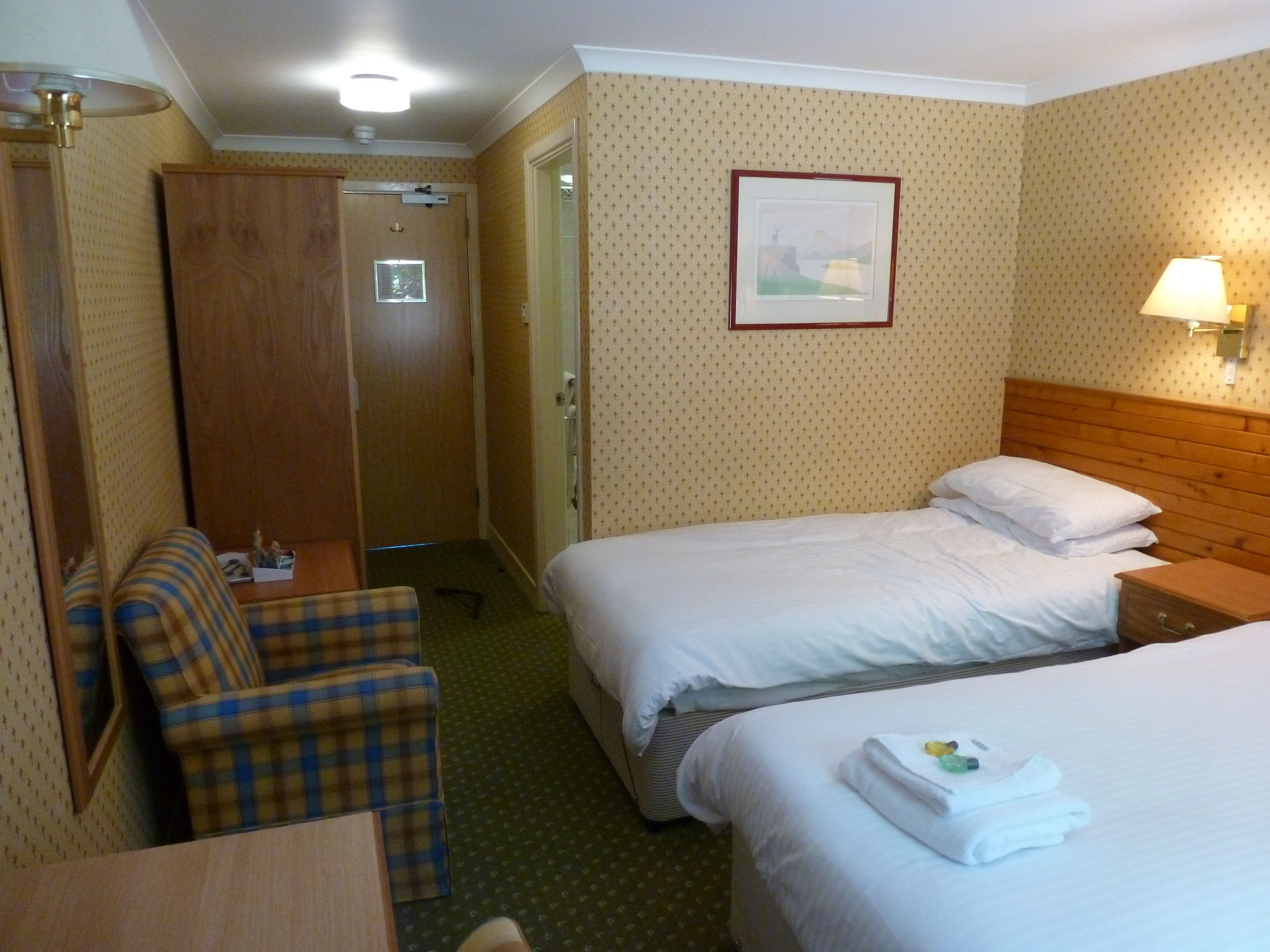 A hotel room with two beds, a wooden headboard, a small table, a chair, and a door.