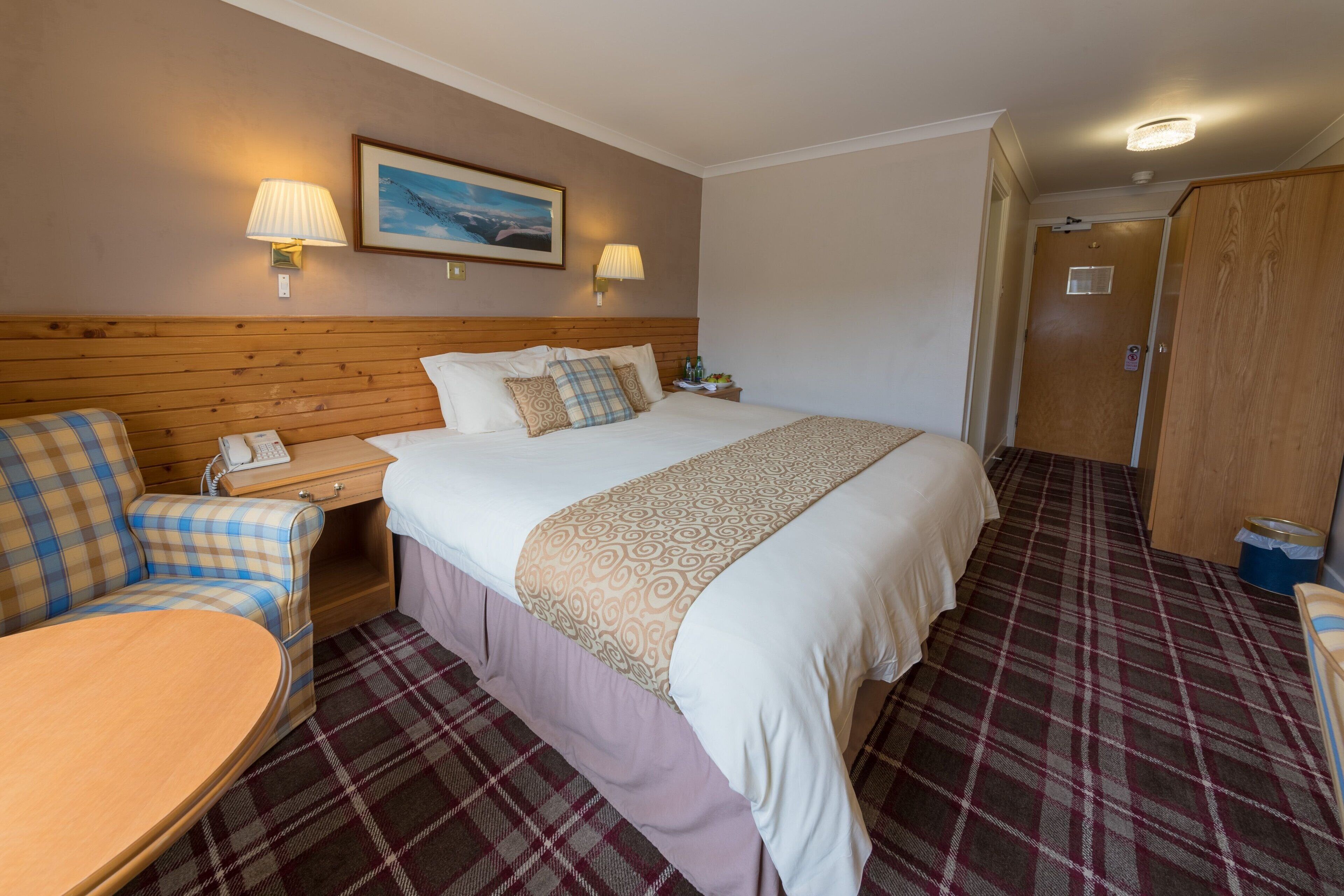 A hotel room with a bed, a chair, a small table, a nightstand, a lamp, and a door.