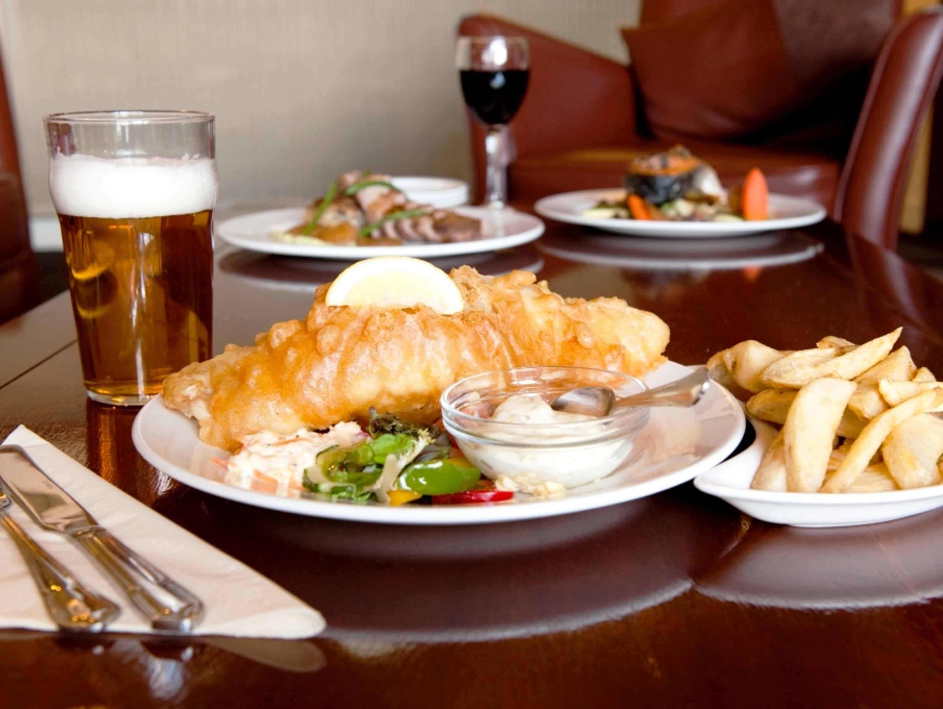 A dining table with a plate of fish and chips, a glass of beer, and a bowl of sauce.
