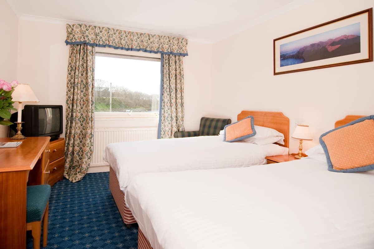 A hotel room with two beds, a desk with a television, a window with patterned curtains, and a framed picture on the wall.