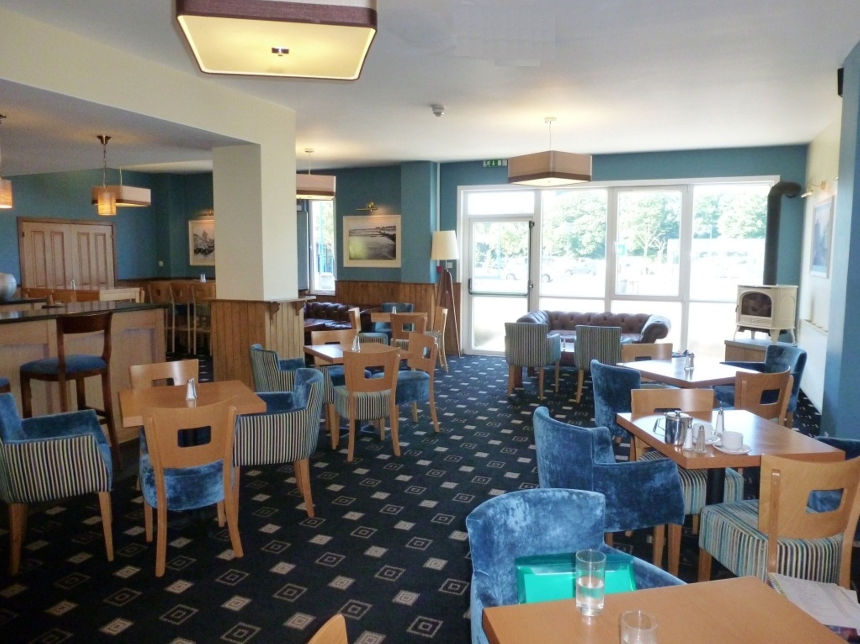 A restaurant with a high ceiling, blue walls, and a patterned floor. There are multiple tables and chairs, and a large window provides natural light.