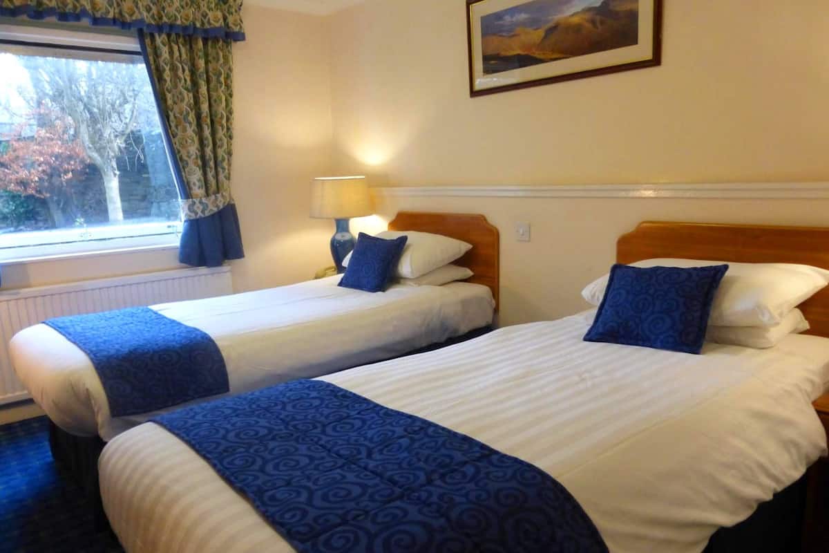 A hotel room with two single beds, each with a blue patterned bedspread and pillows. There is a window with curtains, a lamp on one side, and a framed picture on the wall.