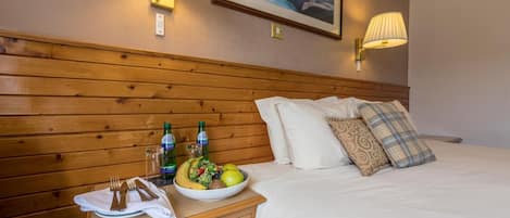 A hotel room with a wooden headboard, a bed with white linens, a bedside table with a bowl of fruit and drinks, and two wall-mounted lamps.
