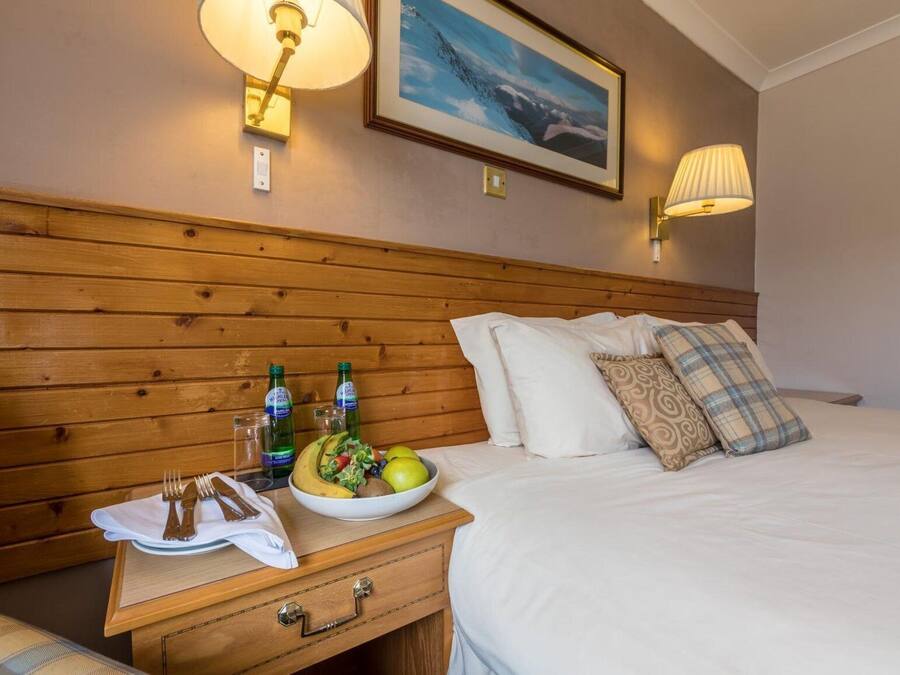 Una habitación de hotel con cabecera de madera, cama con ropa blanca, mesita de noche con un frutero y bebidas, y dos lámparas fijadas a la pared.
