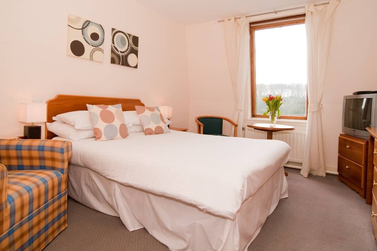 A hotel room with a bed, a TV, a chair, a small table, and artwork on the wall.