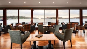 A spacious indoor seating area with multiple tables and chairs, large windows offering a view of the sea and distant hills.