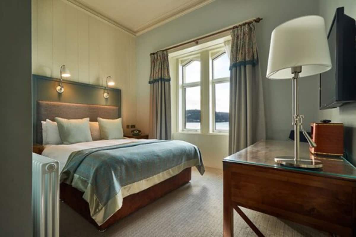 A bedroom with a bed, bedside lamps, a desk with a lamp, a window with curtains, and a view outside.