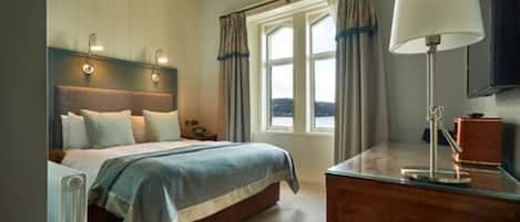 Een slaapkamer met een bed, bedlampen, een bureau met lamp, een raam met gordijnen en uitzicht.