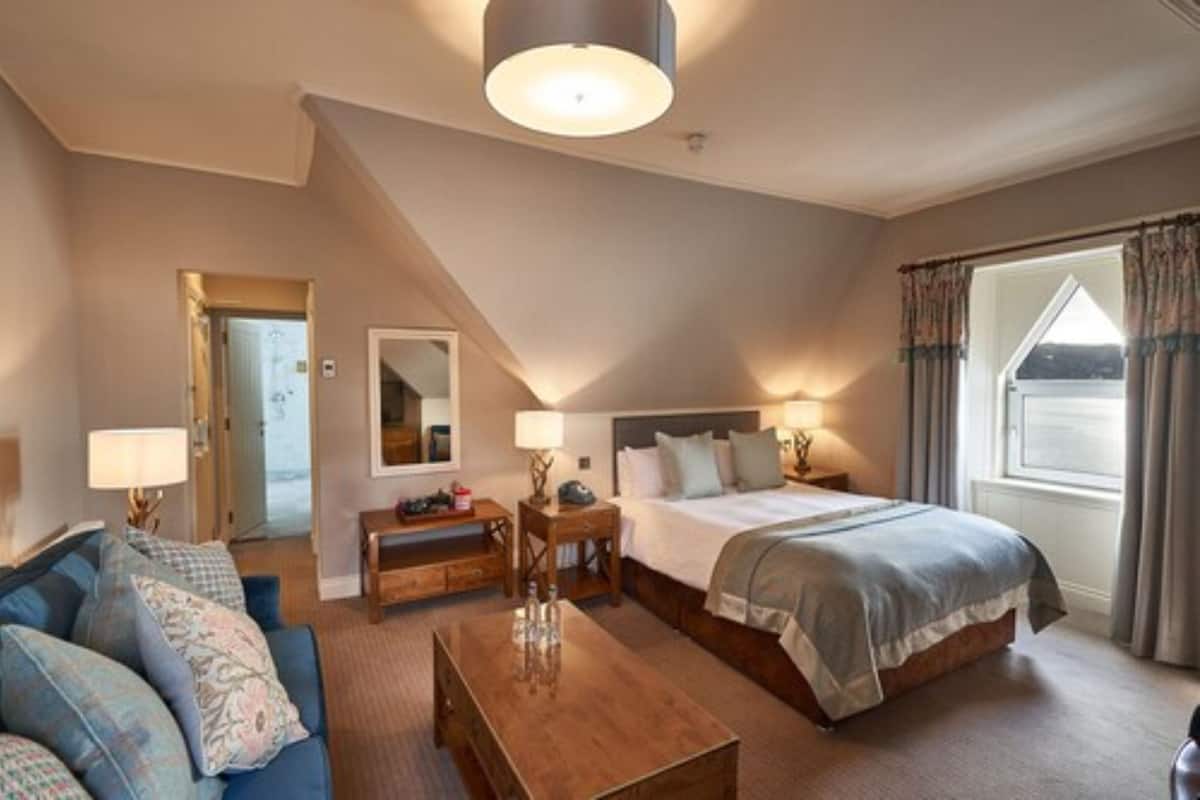 A hotel room with a bed, a sofa, a wooden coffee table, a nightstand, and a lamp. There is a window with curtains and a view of the outside.