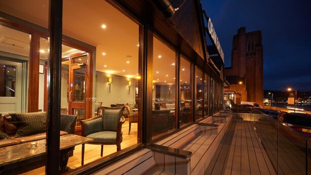 A well-lit outdoor seating area with wooden benches and chairs, large windows, and a view of the cityscape at night.