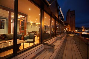Un área de estar al aire libre bien iluminada, con bancos y sillas de madera, ventanas amplias y vista panorámica de la ciudad de noche.