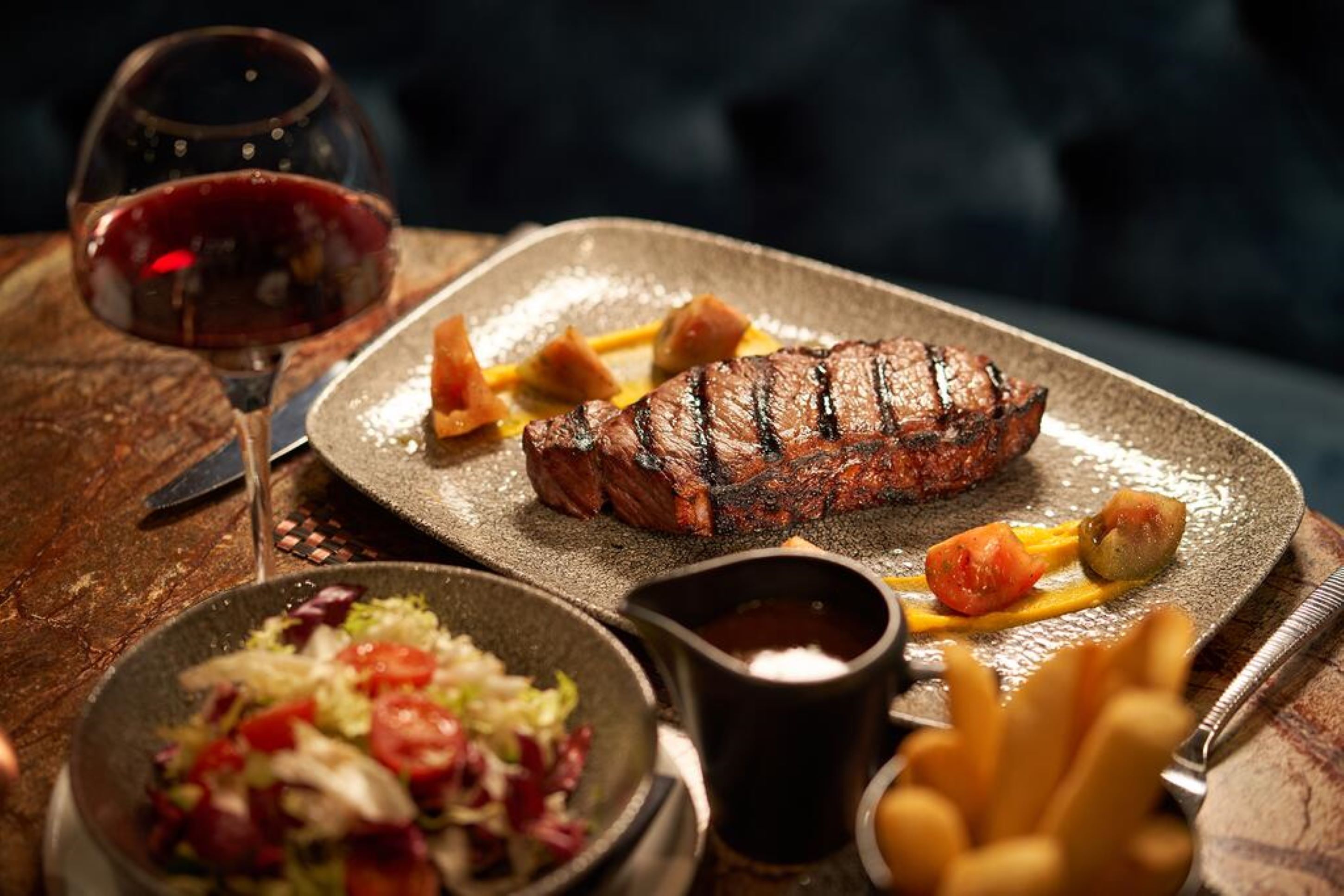 A steak with fries and a salad on a plate, a glass of red wine, and a fork on a wooden table.