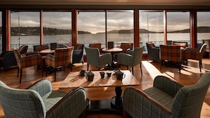 A spacious lounge area with a large table and chairs, large windows offering a view of a body of water and distant hills.