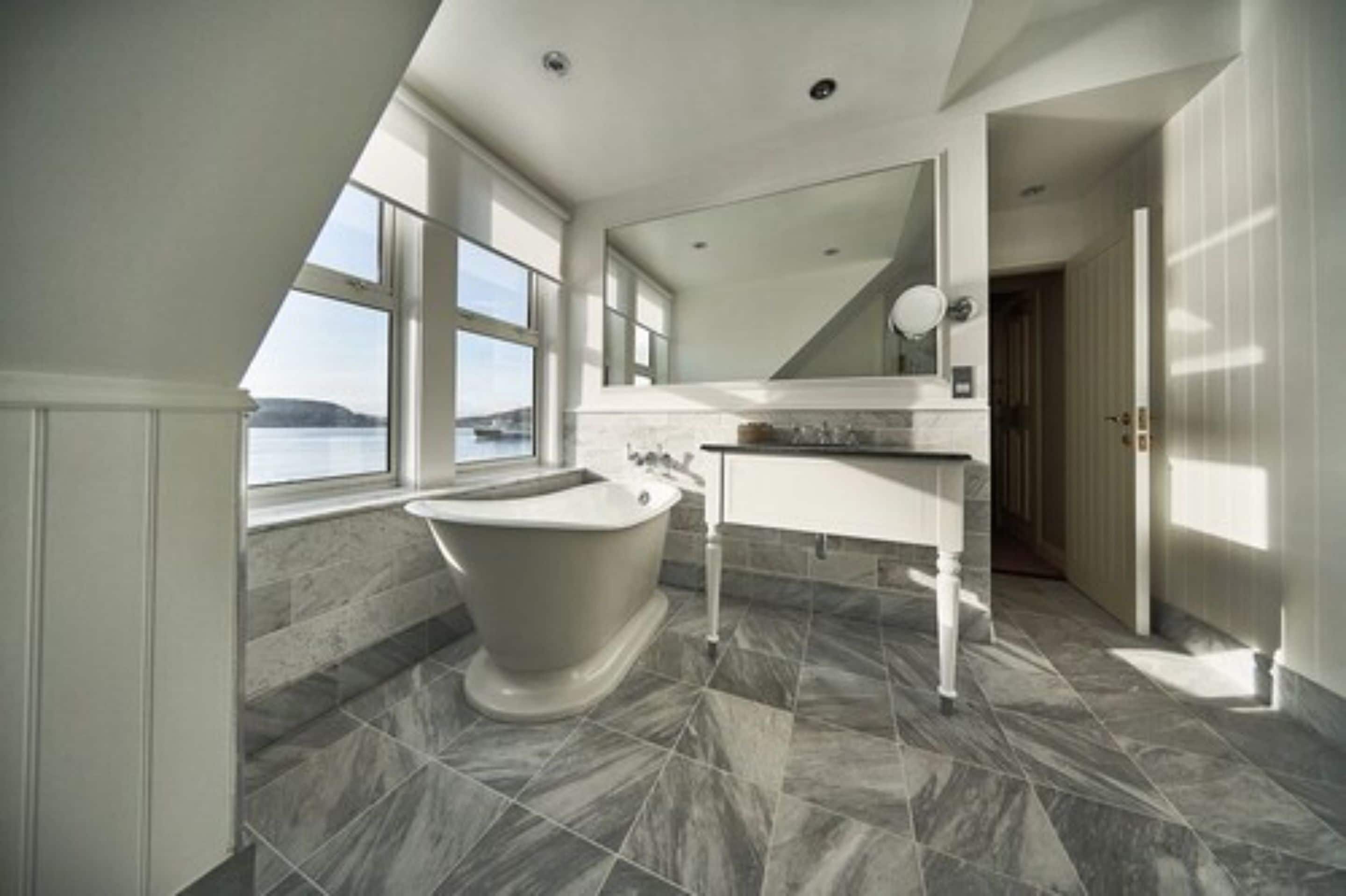 A modern bathroom with a freestanding bathtub, a large mirror, and a view of the sea through large windows.