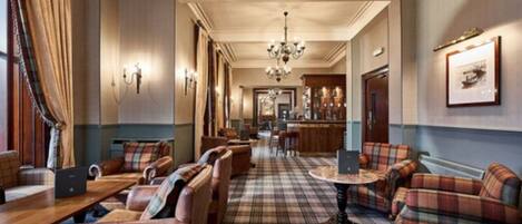 A hotel lobby with a checkered floor, leather seating, a bar area, and a chandelier.