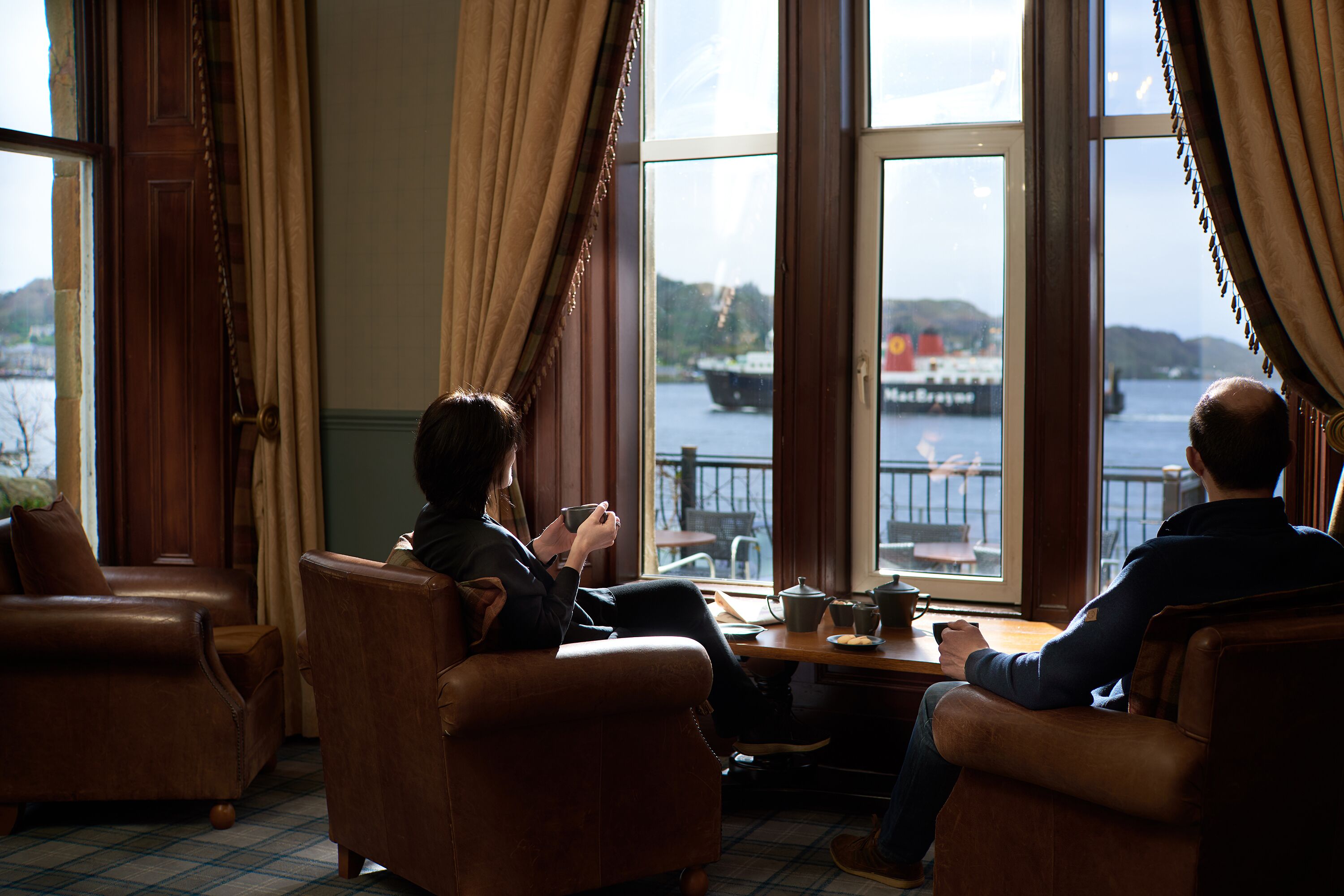 Ein Zimmer mit zwei Ledersesseln, einem Holztisch und Blick auf ein Gewässer mit einem Schiff.