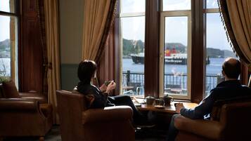 Ein Zimmer mit zwei Ledersesseln, einem Holztisch und Blick auf ein Gewässer mit einem Schiff.